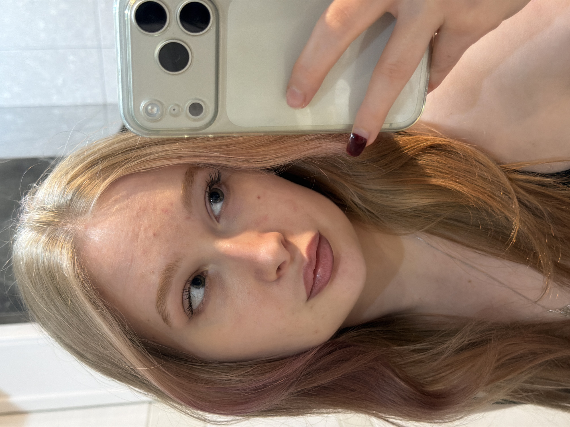 A person is holding a smartphone with multiple camera lenses in front of their face, presumably taking a selfie. The person has fair skin, light brown hair with pink highlights, and is wearing lipstick. The background appears to be a white wall, possibly in a bathroom, with faint dark lines or patterns visible. The image is rotated, with the person's face and the phone angled to the left. There is not enough context to identify the specific location in Rossiyskiy, Russia.