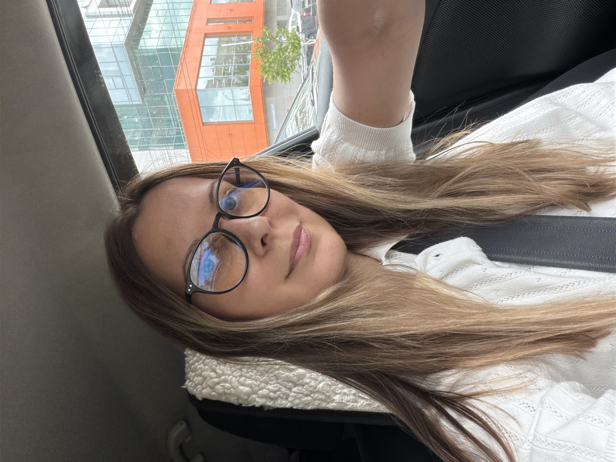 A selfie is taken by a woman in a car, with a view of an urban streetscape through the car window. The woman, with long blonde-brown hair and wearing glasses and a white top, is positioned in the foreground, facing the camera. Behind her, the car's interior is visible, including a seatbelt and a fluffy white material on the seat. Through the window, modern buildings with glass facades and an orange-colored structure are prominent. The background suggests a commercial or business district in Izhevsk, Russia, indicated by a street sign with Russian text. The lighting suggests it is daytime.
