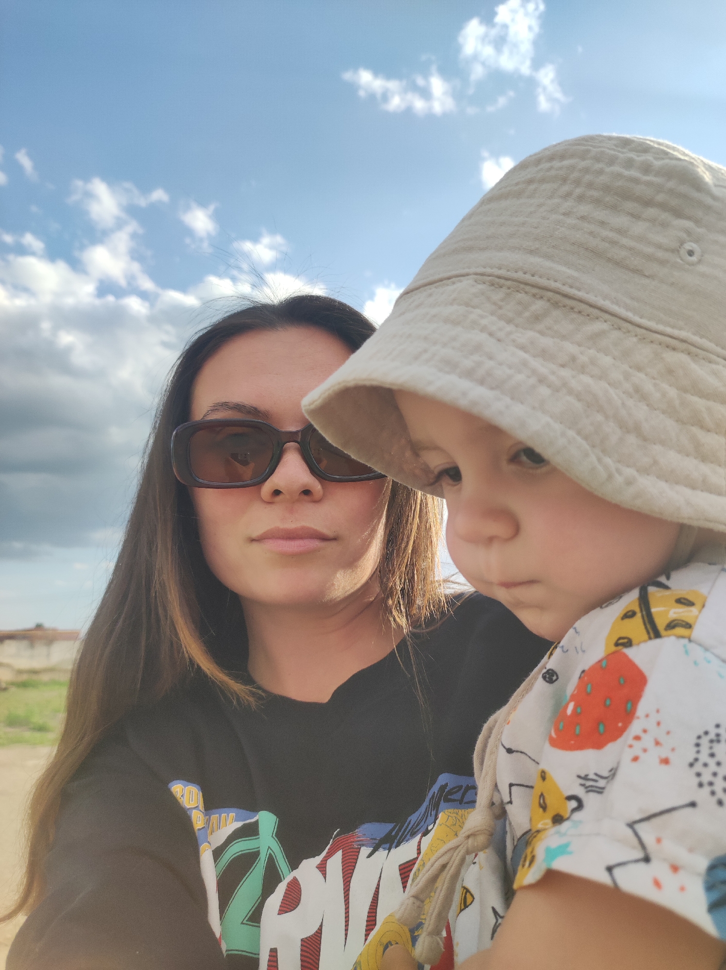 A woman wearing sunglasses and a black shirt with colorful graphics is holding a young child wearing a white bucket hat and a light-colored shirt with colorful designs. The background shows a cloudy sky and a somewhat barren landscape with a small building or structure visible in the distance. This photo was taken somewhere near the city of Chaikovsky, Russia.