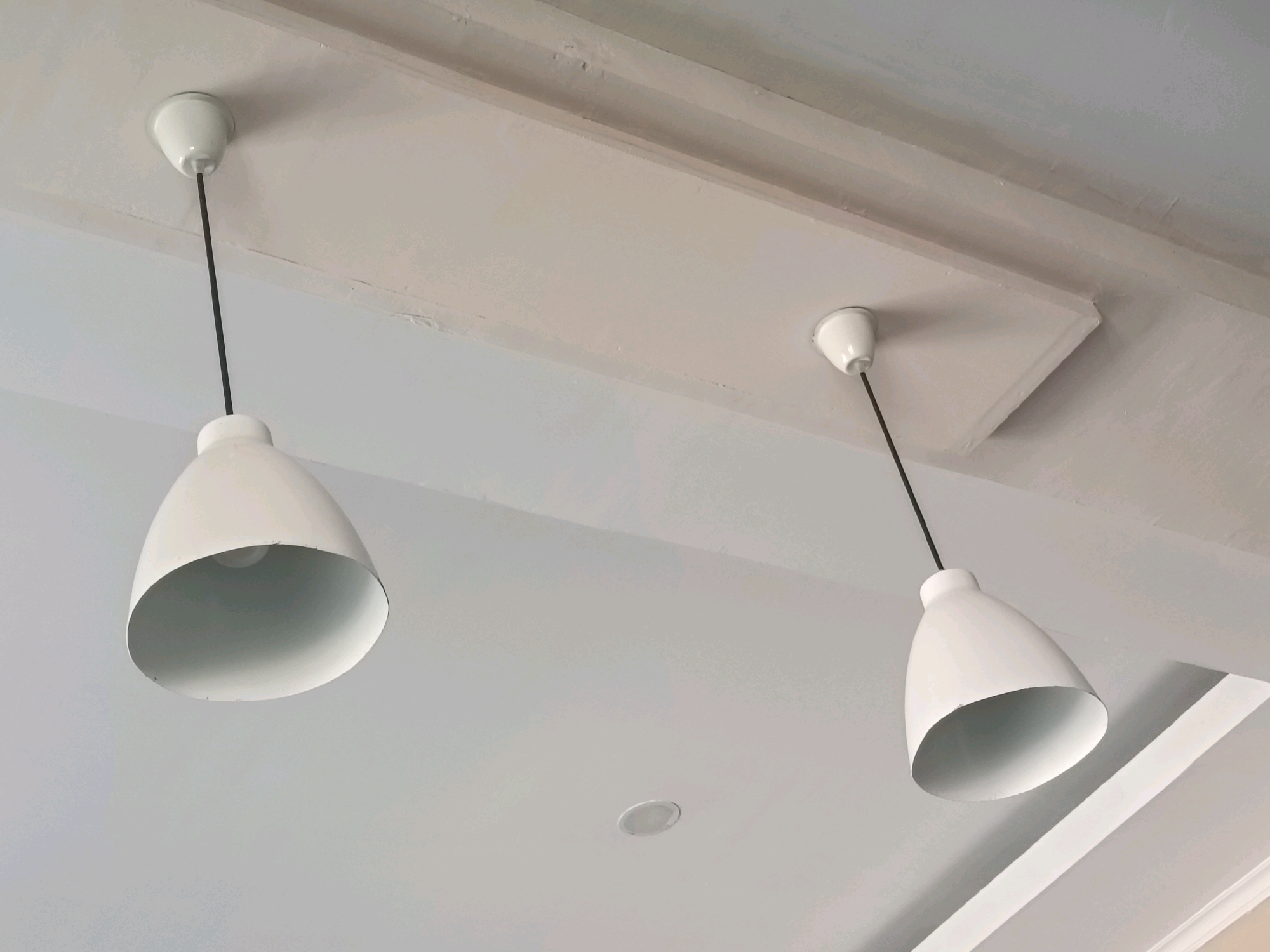 The image shows two white pendant lamps hanging from a white ceiling. The lamps have a conical shape and a black cord connects them to the ceiling. One lamp is on the left side of the image and the other is on the right. The ceiling appears to be made of plaster and has some textured details. There is also a recessed light fixture visible in the center of the ceiling. The lighting suggests an indoor environment, possibly a room or hallway. The scene is static and no people or actions are depicted. The location is not specified in the image.