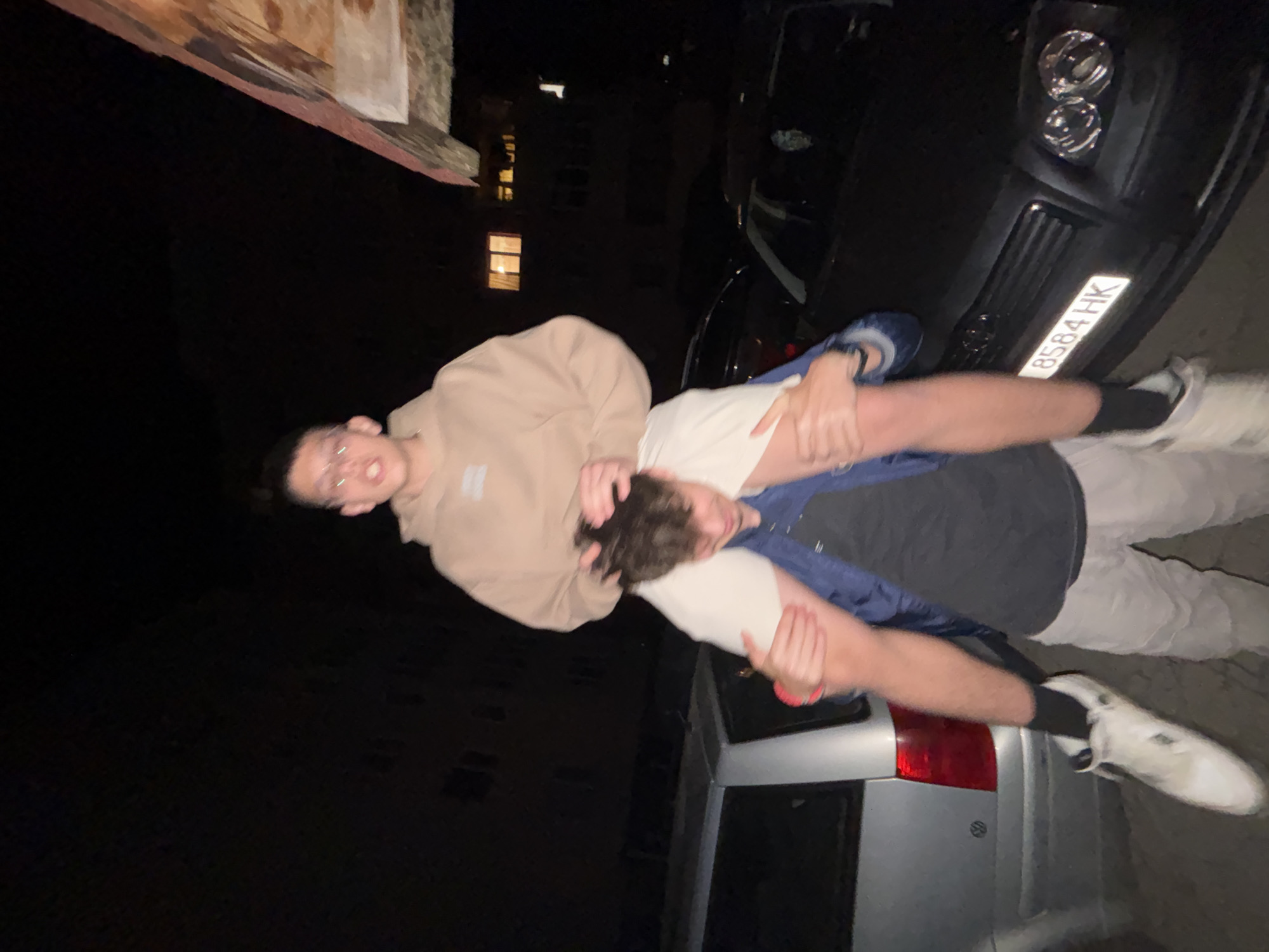 Two young men are photographed at night. One man is carrying the other on his shoulders, with the man on top wearing glasses and a light brown hoodie. The man being carried is wearing a grey t-shirt and blue jacket. Both men are wearing white sneakers. In the background there are two cars, one a dark sedan with a license plate of 8584 HK and the other a silver van. A building with a lit window is partially visible in the background. The location is Troyan, Bulgaria.
