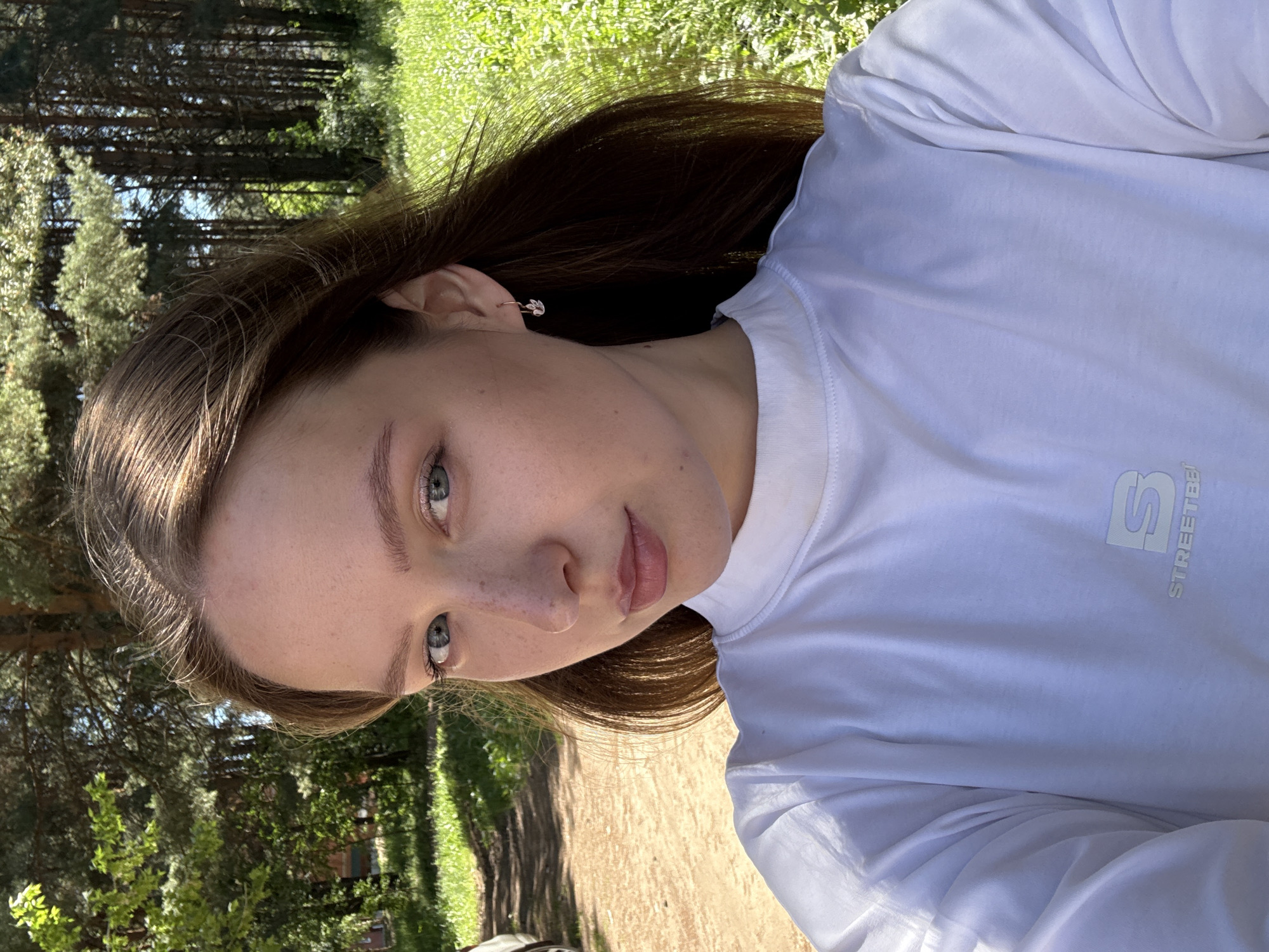 A young woman with light brown hair and blue eyes is taking a selfie outdoors. She is wearing a white t-shirt with a logo that reads "STREETBE". The background shows a path, green grass, and trees. The scene appears to be a park or natural area. The lighting suggests it is daytime. There is no indication of the specific location within Chaikovsky, Russia, other than it being outdoors.