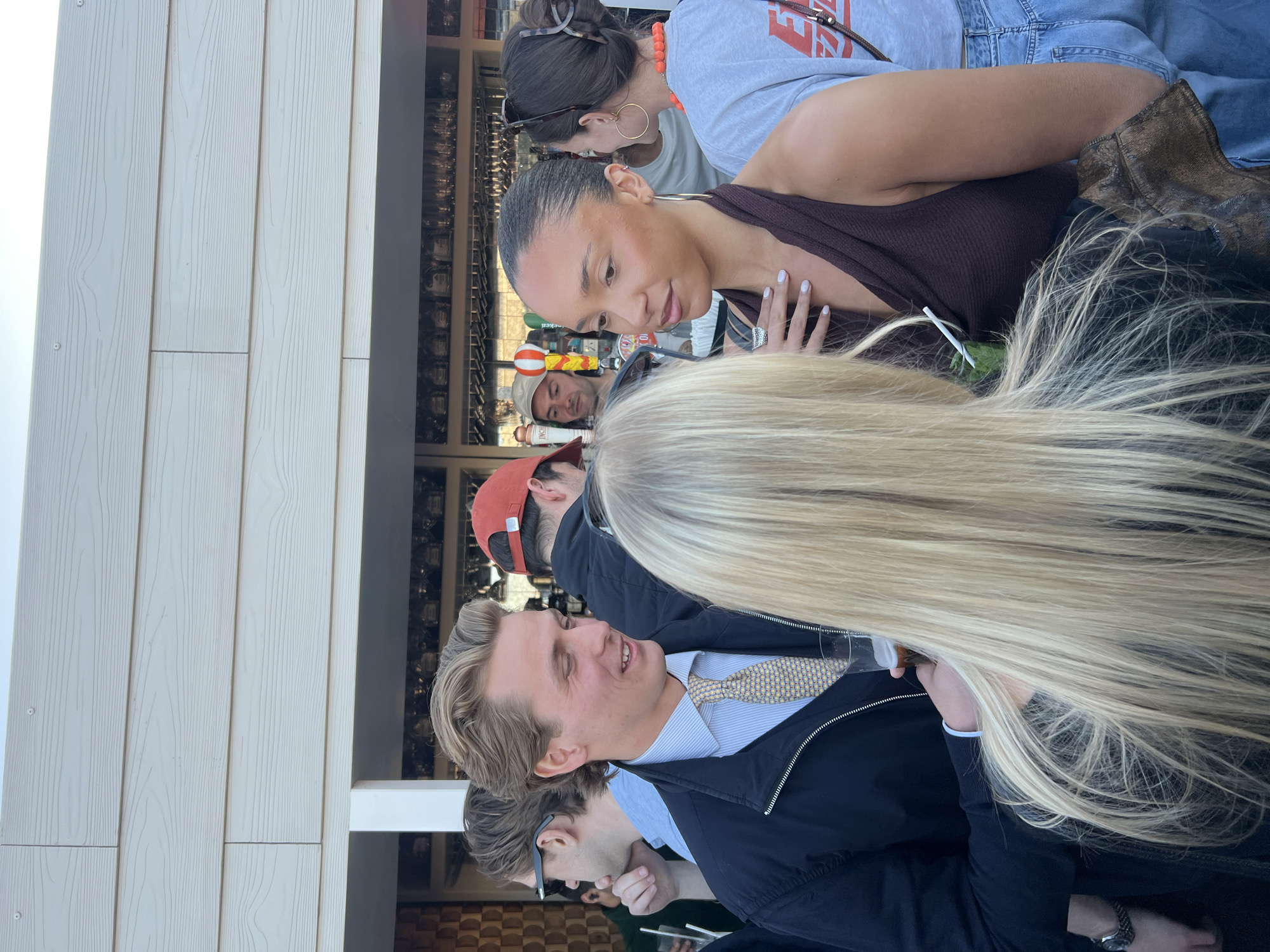 A group of people are clustered together, some interacting and others looking elsewhere. A woman with tan skin and dark hair is wearing a brown tank top and looking down with her hand near her chest. Behind her is a person with an orange necklace and sunglasses perched on their head. A man with blond hair, wearing a blue shirt, patterned tie, and navy jacket, is smiling and looking at someone out of frame. The back of a person with long blond hair is in the foreground, partially obscuring the view. A man in a red hat is looking forward in the background. The setting appears to be an outdoor event or gathering, possibly a bar or restaurant, based on the interior visible in the background. It is not possible to tell if the setting is in London.
