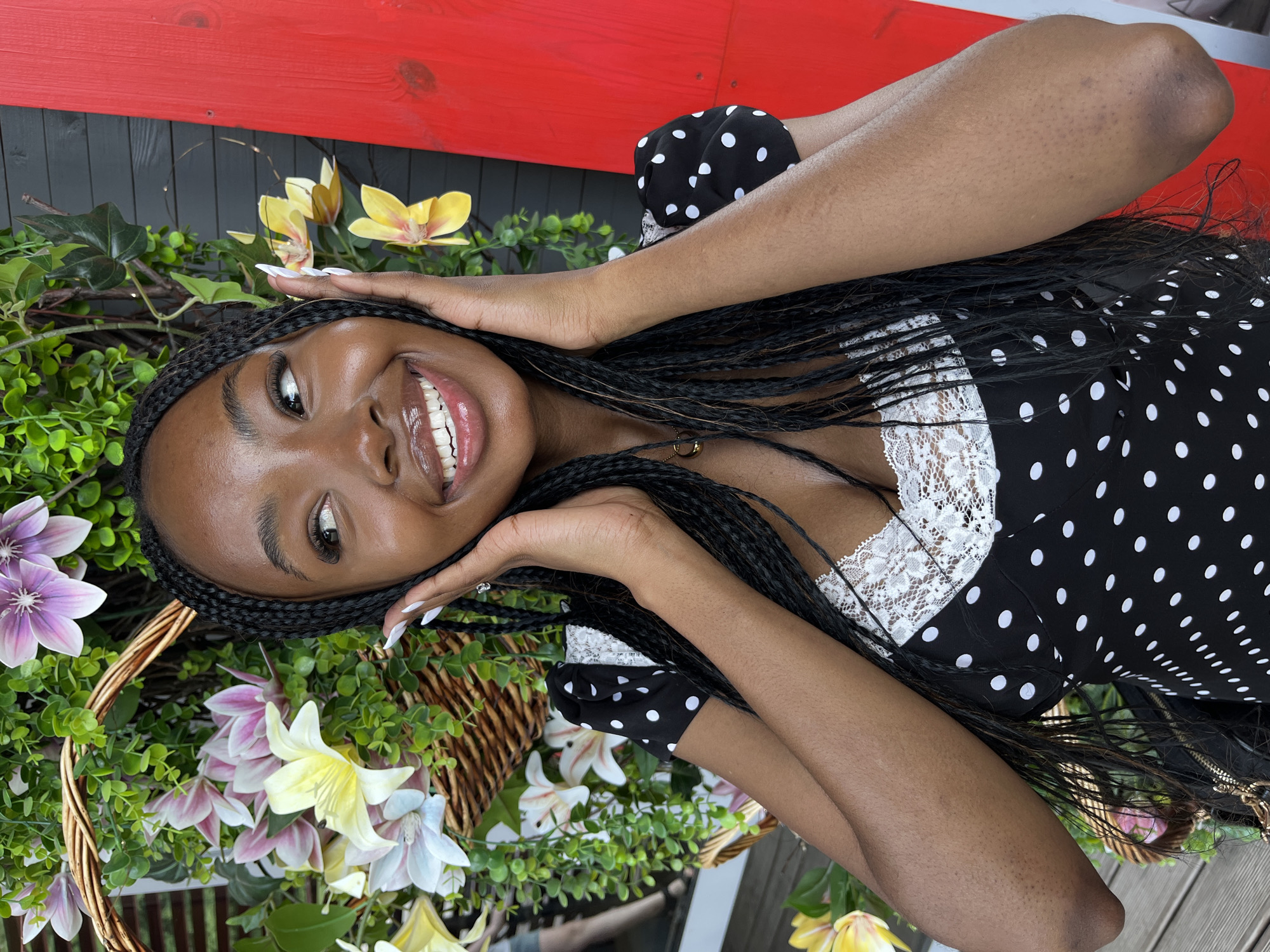 A young Black woman with long braided hair smiles broadly, her hands resting on her cheeks. She is wearing a black dress with white polka dots and a white lace trim. She is positioned in front of a large floral arrangement with various colorful flowers and greenery, set against a dark, textured wall with a bright red vertical element on the right. The ground beneath her appears to be wooden planks. There is no indication of the city of Kazan, Russia, in this image.