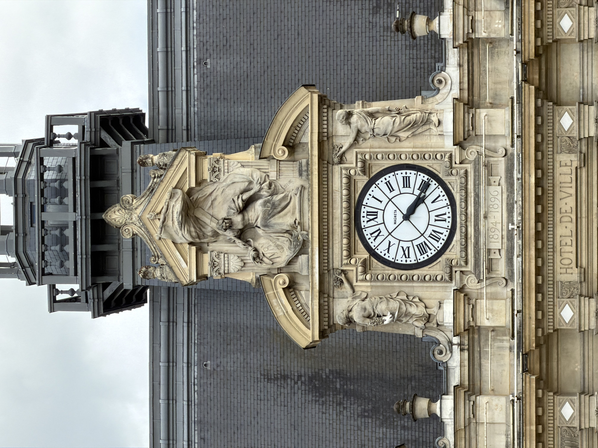 This media file shows a close-up of a clock tower on the exterior of a building. The clock face is round with Roman numerals and black hands. The clock is adorned with stone carvings of figures and decorative architectural elements. Text visible on the building includes "1894-1896" and "HOTEL-DE-VILLE", indicating the building is the town hall constructed between those years. The scene is set against a backdrop of a cloudy sky and a dark brick roof. The overall impression is one of historical grandeur and architectural detail.