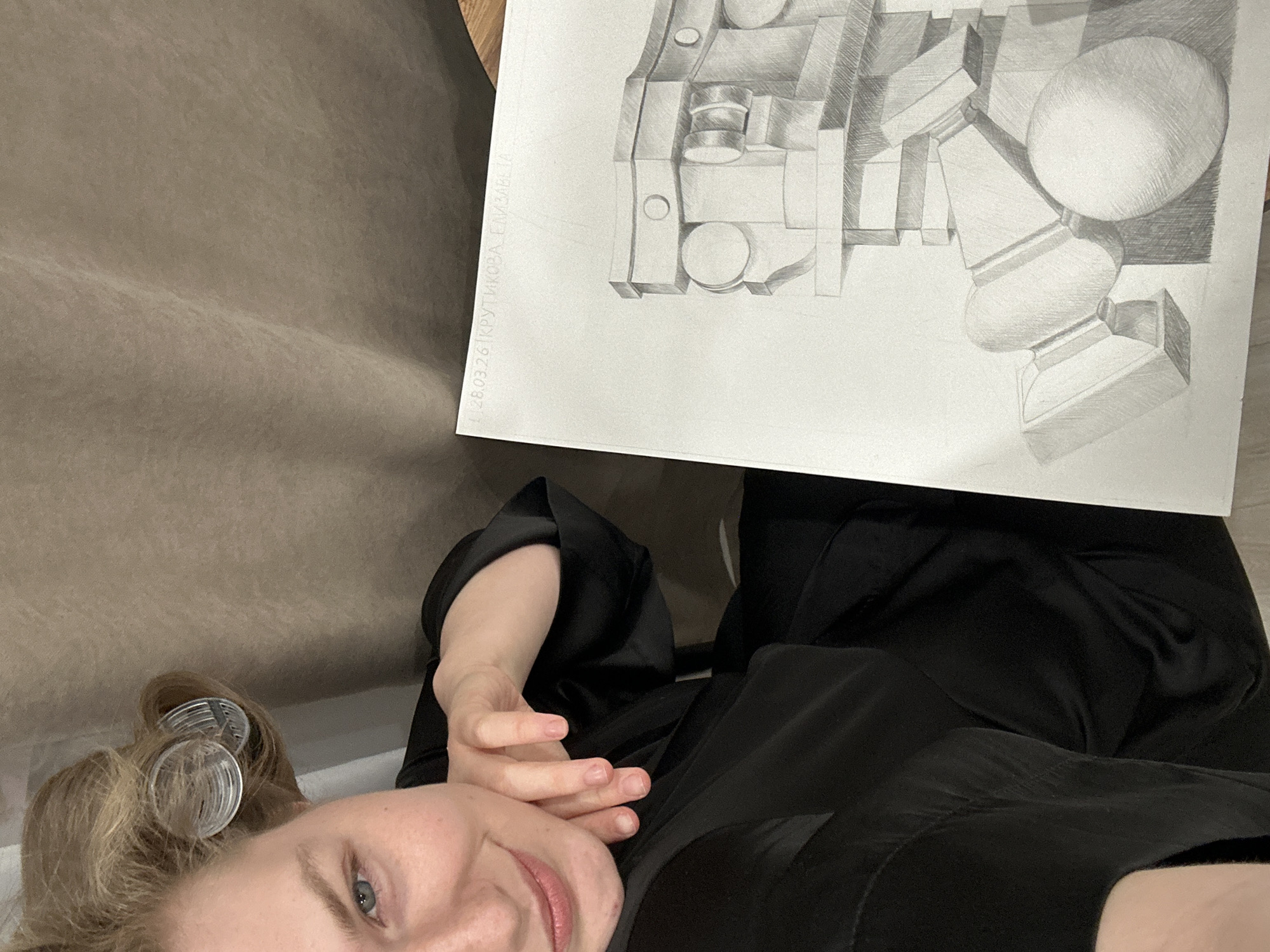 The image shows a woman with blonde hair and hair rollers lying down, holding a drawing in front of her. The drawing is a pencil sketch of geometric shapes, including a sphere, and has some text in Russian along the left edge, possibly a signature and date: "L 28.03.26 | KPYTHKOBA TA". The woman is wearing a black robe or garment. The background appears to be a plain wall or fabric. There is no visible information suggesting the location is Kazan, Russia.