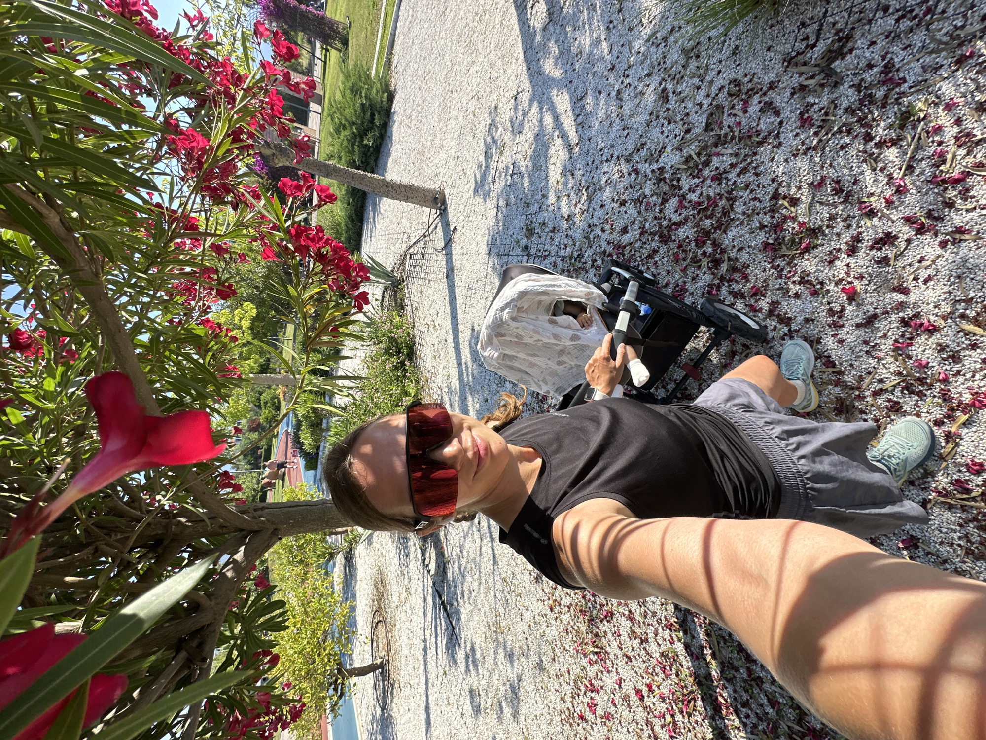 The image is a rotated selfie captured outdoors on a bright, sunny day, depicting a woman pushing a stroller along a gravel path. The woman, who is the primary subject, takes the selfie with an extended arm, her face largely obscured by large, rectangular red-tinted sunglasses. She wears a black short-sleeved top and grey shorts, with light blue athletic shoes visible on her feet.
Beside her, a black stroller is visible, containing a baby or small child partially covered by a white, patterned blanket. The path underfoot is covered in light-colored gravel, extensively scattered with fallen red petals and some dried leaves, creating a decorative pattern. Strong shadows cast by nearby trees and structures indicate bright, direct sunlight, suggesting a midday or early afternoon setting.
To the left of the woman, a lush bush with vibrant red flowers, characteristic of oleander, fills a significant portion of the foreground and extends into the background, adding a burst of color. Beyond the gravel path, there are areas of green grass, additional trees, and subtle hints of buildings or fences in the distance. The overall scene conveys a pleasant outdoor walk in a well-maintained garden or park-like setting, consistent with the Mediterranean climate of Fethiye, Turkey. No visible text is present in the image.