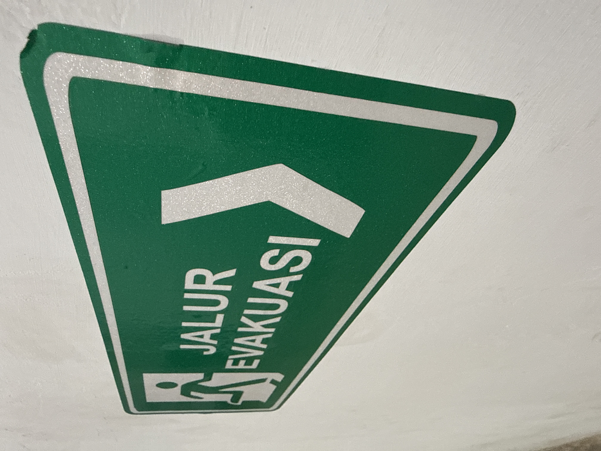 A green and white sign indicating an evacuation route is affixed to a white textured wall. The sign features a silhouette of a person running, a white arrow pointing to the right, and the text "JALUR EVAKUASI" in white capital letters. The sign is positioned at an angle, and the wall appears to be an interior surface. The image was taken in Lhokseumawe, Indonesia.