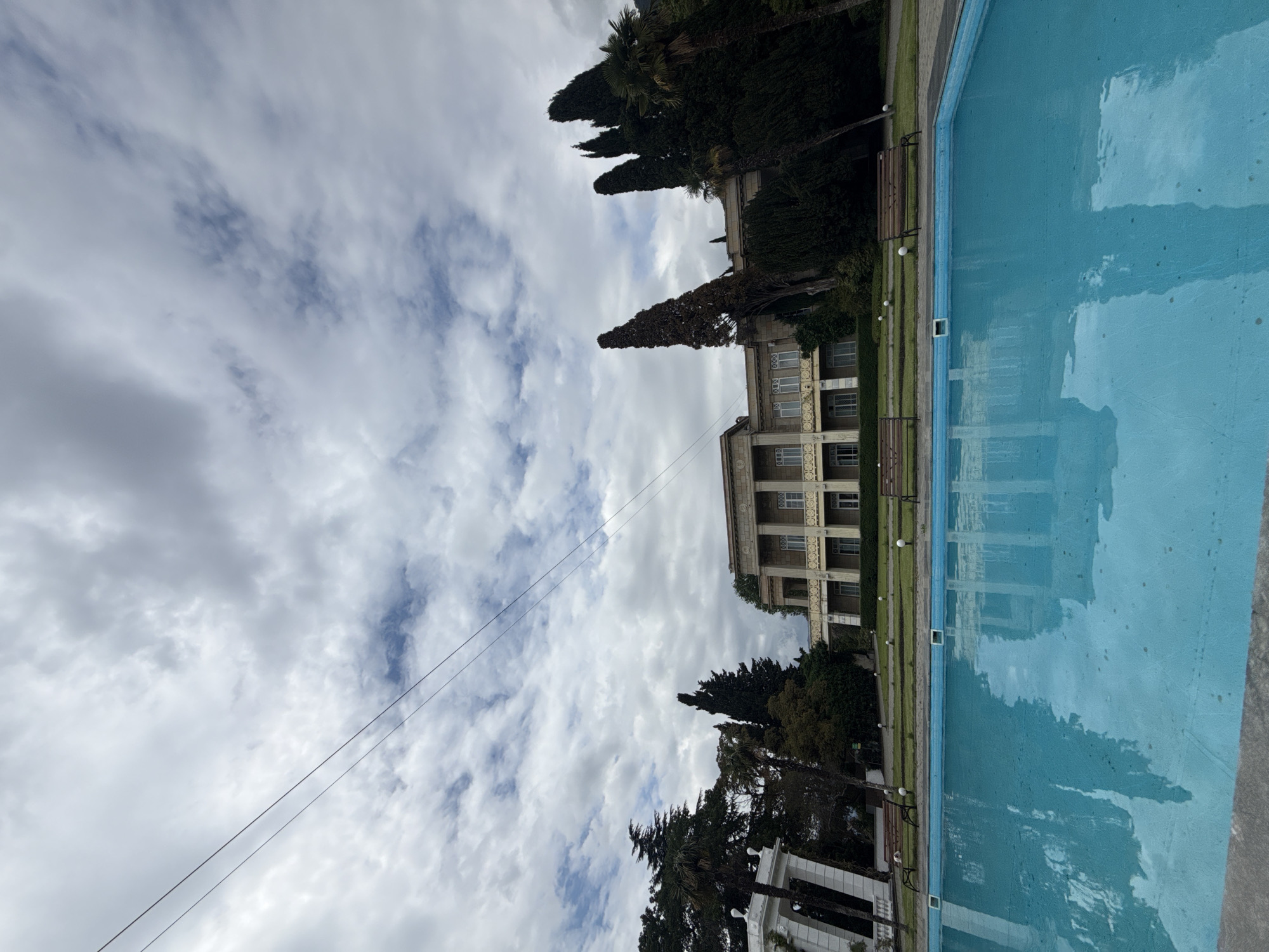 The image depicts a scene in an urban location, likely a park or recreational area. A building with multiple floors and columns is visible, surrounded by tall, dark trees. Beside the building is a rectangular pool filled with blue water, its surface reflecting the sky and surroundings. Above the building and trees, the sky is filled with numerous white clouds. Two power lines cut across the sky. The image is taken from a low angle and is rotated slightly counter-clockwise. No people are visible in the image.