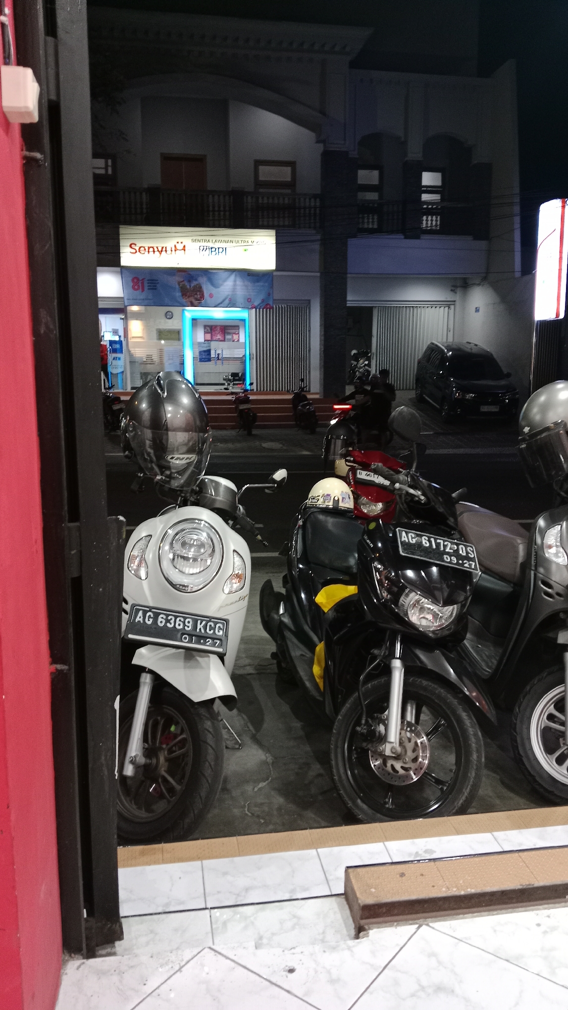 An outdoor, nighttime scene captures the front of a building in Blitar, Indonesia, with several motorcycles parked outside. The building appears to be a commercial establishment, possibly a bank or a service center, indicated by a sign that reads "SENTRA LAYANAN ULTRA V.I.P." and "BPI". An ATM machine is visible inside the building through a glass partition, illuminated by blue neon lights.
In the foreground, three scooters are parked. The leftmost scooter is white with the license plate "AG 6369 KCC" and the date "01-27". A large, dark grey helmet with a reflective visor is placed on its handlebars. Adjacent to it is a black scooter with the license plate "AG 6172 QS" and the date "09-27". A yellow cloth is visible on the seat of the black scooter, and a white helmet with a light-colored visor is placed on top of it. The third scooter, to the right, is mostly obscured but appears to be dark grey, with a silver helmet on its handlebars.
In the background, a black car is parked further back. The building facade is a mix of light-colored walls and dark columns, with windows and balconies visible on the upper floors. A tall, vertical light pole with red accents stands on the right side of the frame. The ground is a mix of concrete and tiled surfaces. The overall impression is of a well-lit commercial area at night, with motorcycles serving as a common mode of transportation.