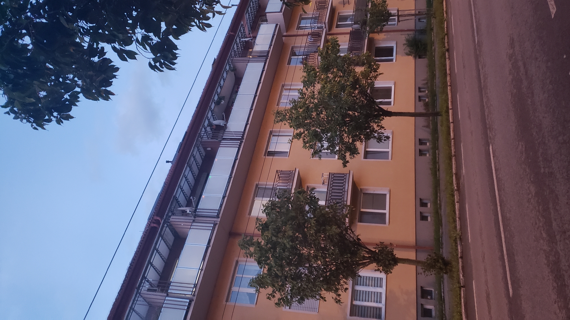The image captures a street-level view of a light orange, multi-story apartment building in Prešov, Slovakia, under a twilight sky. The building features multiple floors with balconies that have glass railings and metal supports. Various windows, some with blinds, are visible on the facade. In front of the building, several trees with green foliage are planted along a grassy strip that separates the building from the asphalt road. The road has white dashed lines marking lanes. The overall scene suggests a residential area during dusk.