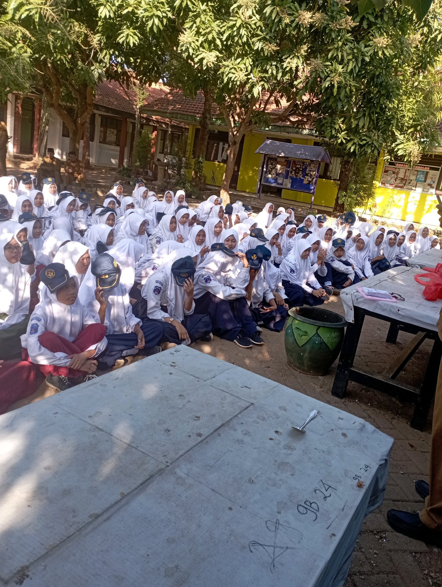 In Banyuurip, Indonesia, a group of students, likely at a school given the uniforms and setting, are gathered outdoors under trees, sitting on the ground. Most are wearing white hijabs and uniforms, suggesting a Muslim educational institution. The students are facing forward, with some appearing to listen or observe something out of frame. Several individuals in the front row are more distinctly visible, including two students wearing dark caps with logos. One of these students is in the foreground, sitting cross-legged, wearing a maroon skirt and white shirt. Another student next to them wears a dark blue skirt and white shirt. Behind them, a large collection of students are seated. A rustic wooden table covered with a light-colored cloth is in the foreground, with some writing on it and a spoon. A large, green, decorative pot sits beside the table. In the background, buildings with tiled roofs and trees suggest a school campus or similar outdoor educational facility. The overall scene depicts an outdoor assembly or lesson.