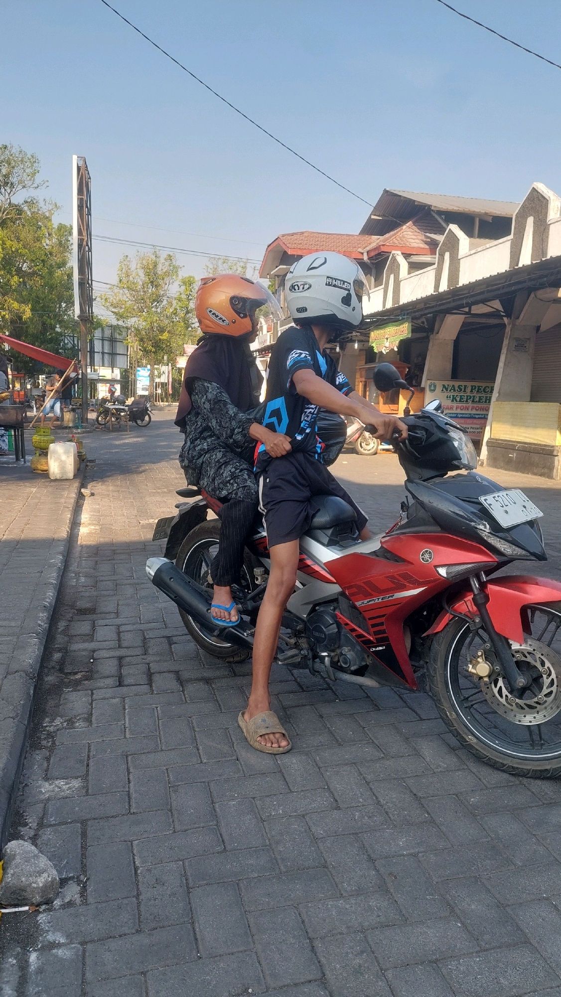 Two individuals are seated on a red and black Yamaha Jupiter motorcycle parked on a brick-paved street in Nologaten, Indonesia. The person in the rear, wearing a dark hijab and an orange helmet, is likely female and is dressed in a patterned black and grey outfit. The person in the front, wearing a white helmet, is dressed in a black t-shirt with blue accents and black shorts, suggesting they are male and is operating the motorcycle. Both are wearing helmets, and the rear passenger has blue flip-flops on. The background shows a building with arched windows and a sign that reads "NASI PECEP 'KEPLER'." Trees and other vehicles are visible in the distance. The lighting indicates it is daytime.