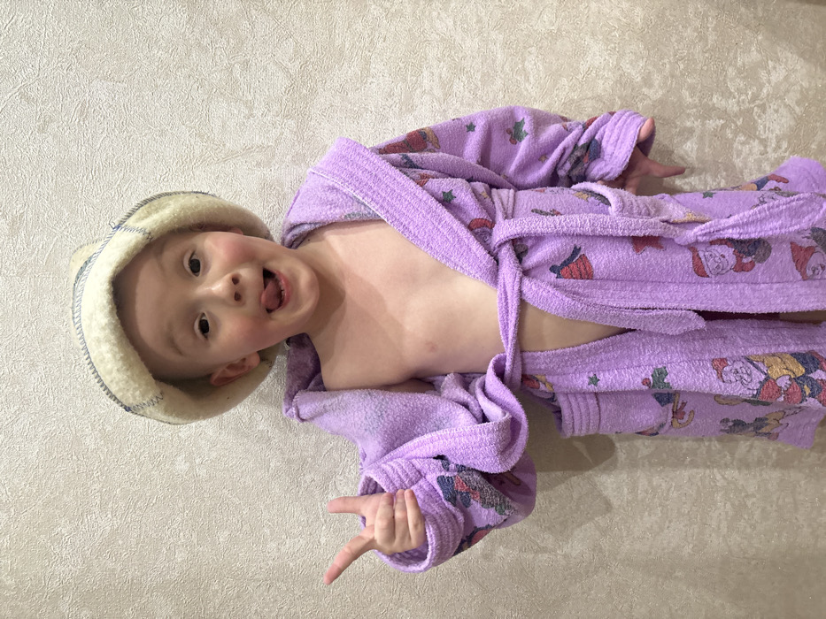 An indoor full-frame photograph depicts a young child, approximately 3-5 years old, positioned centrally against a light beige, textured wall in Pavlovsky, Russia. The child, with fair skin and visible short hair under headwear, wears a light cream-colored, possibly wool, hat with blue stitching, and an oversized, light purple terry cloth bathrobe. The bathrobe is secured at the waist with a tie, leaving a portion of the child's chest exposed. The robe's fabric is patterned with various cartoon characters and holiday-themed motifs including bells, stars, and dwarf-like figures. The child's right hand (viewer's left) is raised to chest height, forming a "V" or peace sign with the index and middle fingers extended. The child's face is directed towards the camera, eyes wide open, with the mouth open and tongue extended in a playful gesture. The lighting is even, casting no significant shadows.