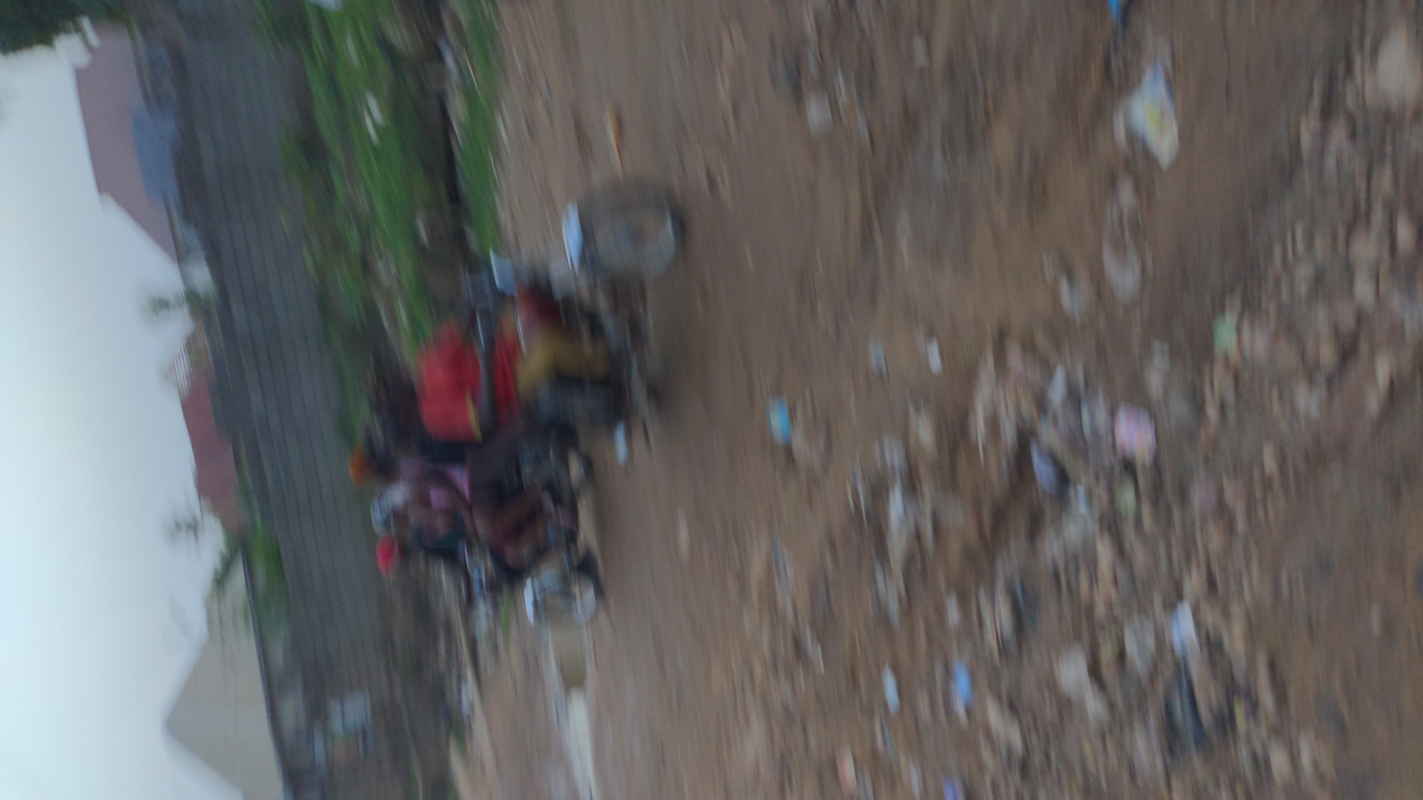 The image shows a scene from Ado, Nigeria. There are multiple people on a motorbike driving down a dirt road. The road is littered with trash. A building and vegetation are visible to the side of the road.