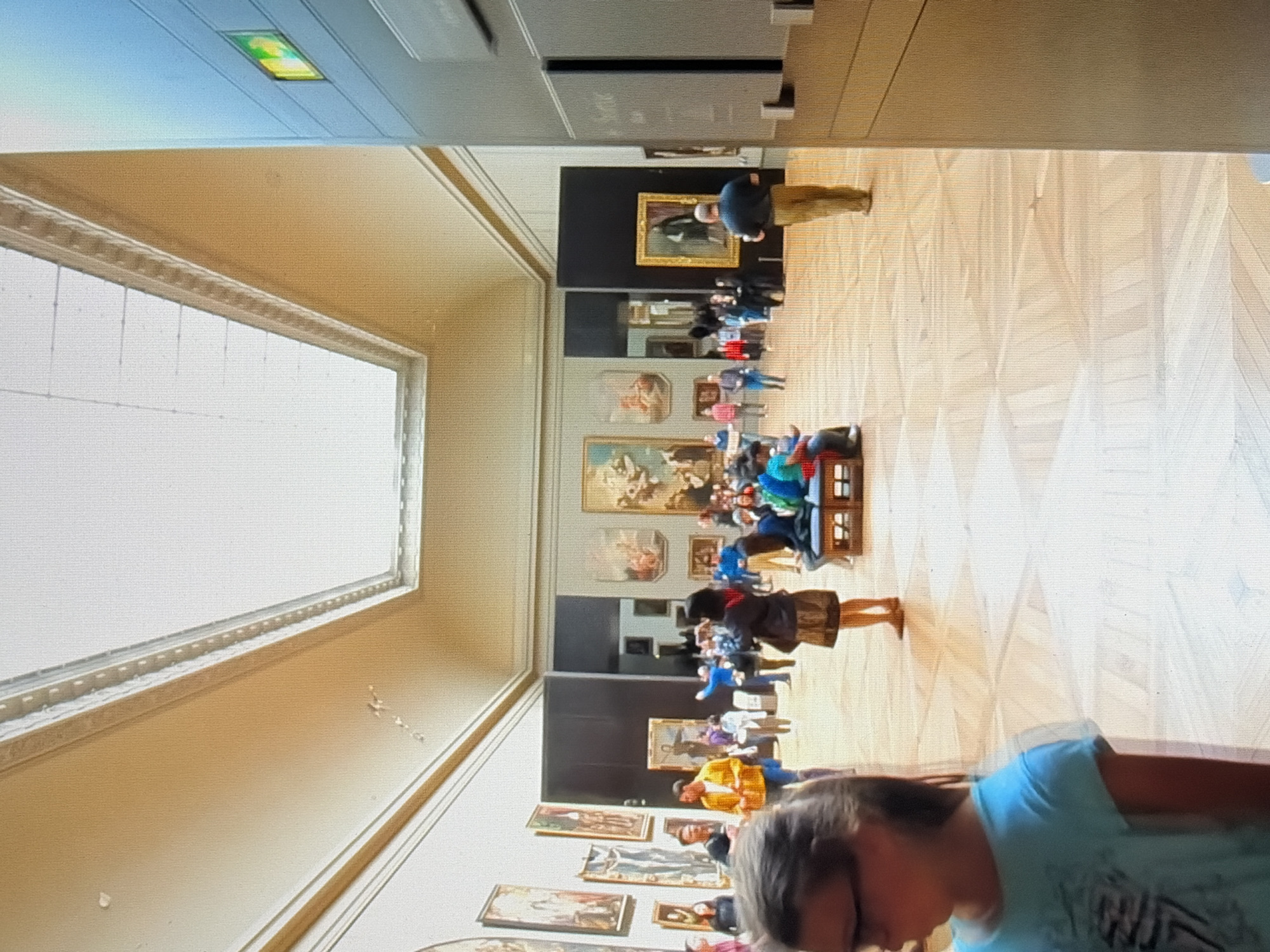 The image depicts an interior view of a museum gallery with a high, arched ceiling and large skylights. The walls are lined with numerous paintings in ornate frames, displayed at various heights. Several groups of people are visible, some standing and observing the art, while others are seated on benches or walking through the expansive space. The floor is made of polished wood with a geometric pattern. In the foreground, on the left, a young girl with glasses and a blue shirt is partially visible. On the right, a man in a dark shirt and khaki pants stands facing away from the camera, looking at a painting. A green emergency exit sign is mounted on a panel on the right side of the frame. The overall atmosphere suggests a busy but orderly museum setting.