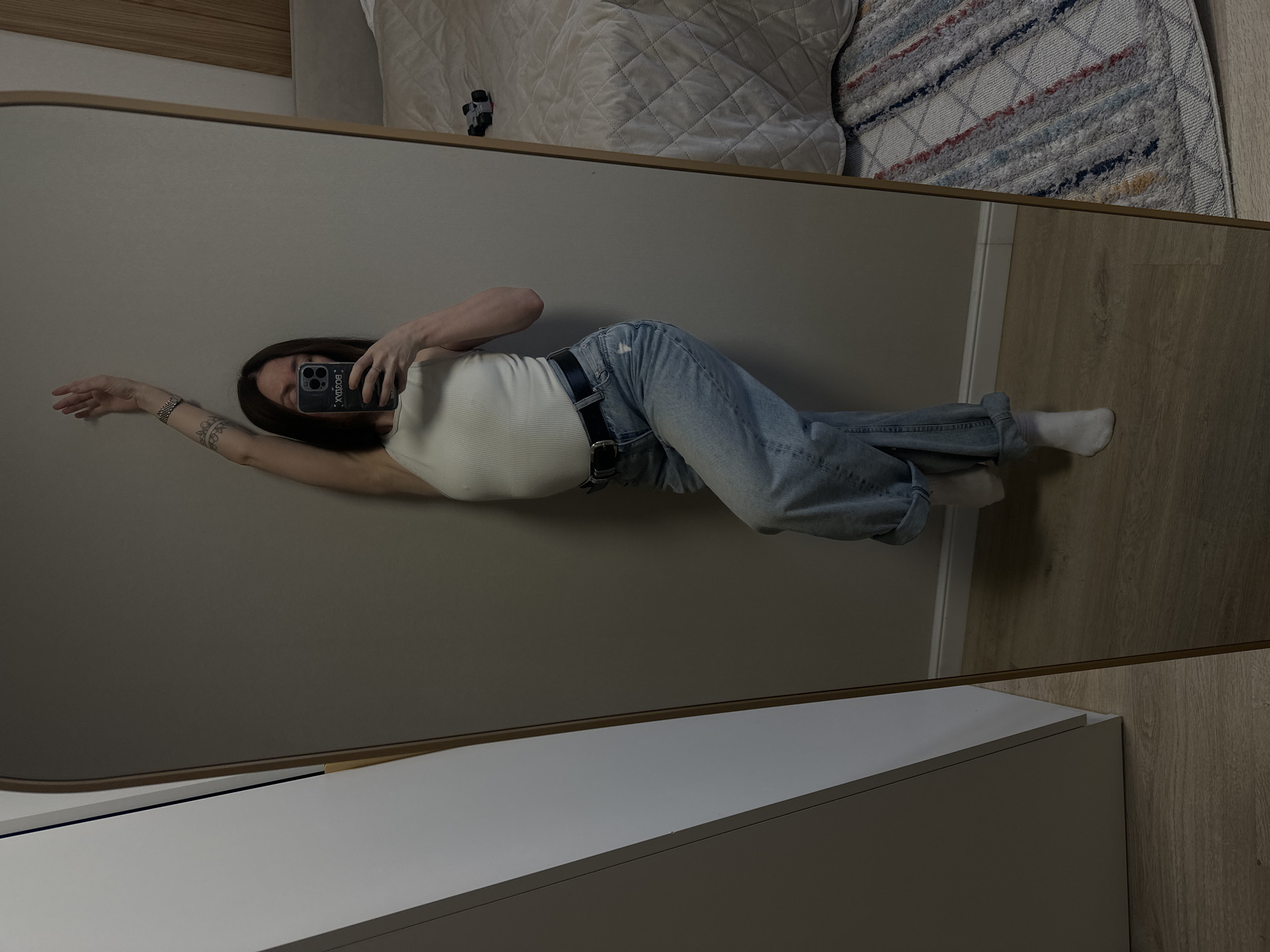 The image captures a mirror selfie of a person in an indoor setting in Votkinsk, Russia. A woman with long, dark hair is reflected in a full-length mirror, lying on her back with her body positioned diagonally. She is wearing a light-colored, ribbed tank top, light blue denim jeans with cuffed ankles, a black belt, and white socks. Her left arm is extended outwards, revealing a visible tattoo on her forearm, and her right hand holds a dark-colored smartphone (possibly an iPhone with multiple cameras) that obscures most of her face.
The phone case features visible mirrored text in Cyrillic, which, when read directly from the reflection, appears to include words such as "УХОДЯ", "ВВОДА", "ВОДА", and "УХОД". The mirror has a thin, light-colored frame.
In the background of the reflection, above the woman's head, is a bed or couch covered with a white quilted blanket or comforter, and a small, dark object rests on it. To the right of the bed, a patterned rug with stripes of various colors (including blue, red, white, gray, and yellow) is visible on a light wood floor. In the foreground, below the mirror, is a white piece of furniture, likely a dresser or cabinet, with a light wood floor extending to its right. The overall setting suggests a casual, residential environment.