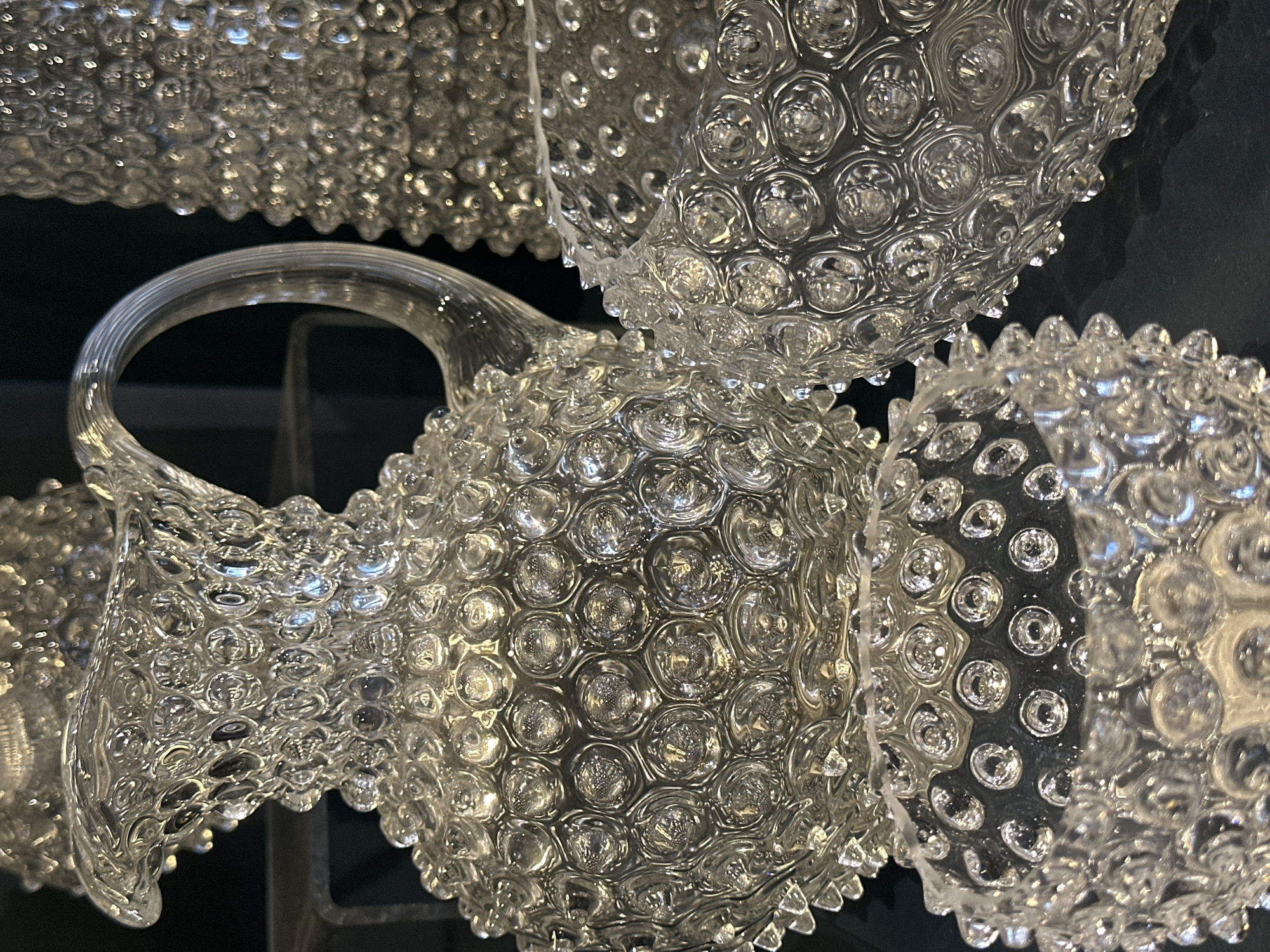 The image shows a close-up of clear glass items, likely decorative bowls or pitchers, featuring a textured surface with raised circular bumps. One item has a handle. The background is dark, possibly a dark wall or surface. The location in Tunbridge Wells, United Kingdom is not apparent from the image.