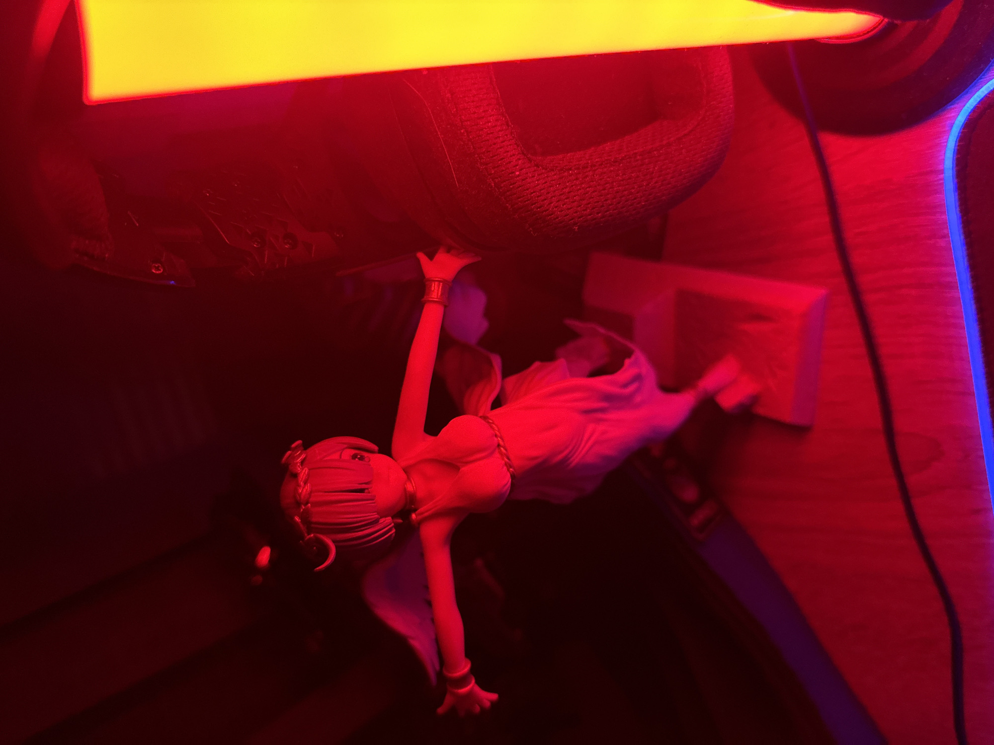 In an indoor setting, a figurine of a winged woman with a halo is positioned on a wooden surface. The figurine is depicted in a light-colored dress and has outstretched arms. To the right of the figurine, a portion of a black textured object is visible, possibly a shoe. A tall, illuminated object with a yellow-to-orange gradient is situated on the far right, casting a bright light. The scene is illuminated by red and blue lighting, creating a dramatic effect. The overall impression is that of a dimly lit room with artificial lighting.