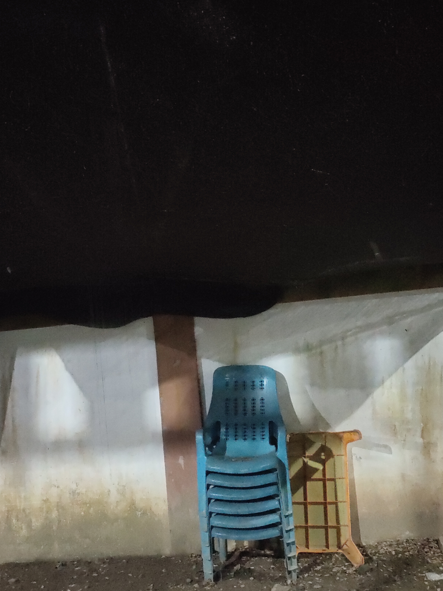 In Keudematangkuli, Indonesia, a stack of blue plastic chairs is positioned against a light-colored wall with a brown stripe. To the right of the chairs, an orange plastic stool with a grid pattern is leaning against the wall. The ground is covered in dark gravel and debris. Above the wall, a dark, textured ceiling is visible, with a slight curve in the middle. The scene appears to be an outdoor or semi-outdoor area, possibly a shelter or covered space.