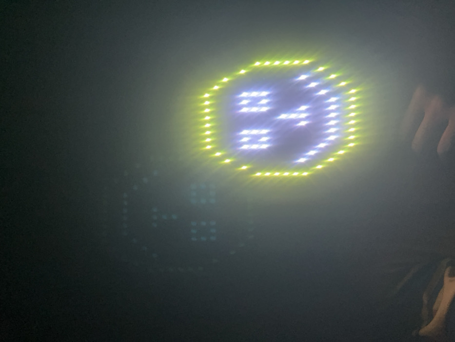 A dark image shows two digital displays. The top one is a glowing yellow and blue circle with a stylized face. Below and slightly to the left is a faded and less illuminated version of the same design. A human hand and a small section of clothing are visible in the right corner. The image has a blurred, foggy appearance, possibly due to low light conditions.