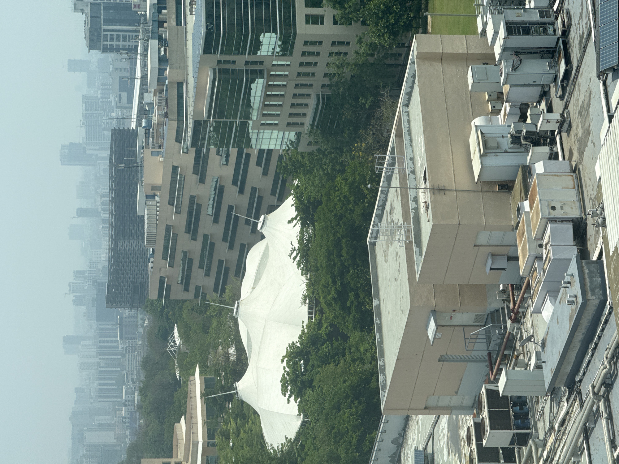 The image depicts a dense urban landscape, possibly in Man, India, given the prompt. In the foreground, a flat rooftop of a building houses numerous industrial air conditioning units and various pipes. Adjacent to this building, a large, white tensile structure, resembling a sunshade or canopy, is nestled amongst dense green trees. Beyond this, taller buildings rise, showcasing modern architectural designs with reflective glass facades. In the far distance, a hazy skyline reveals a multitude of structures, blending into a somewhat obscured horizon, suggesting either atmospheric pollution or significant humidity typical of urban Indian environments. There are no people visible in the image.
