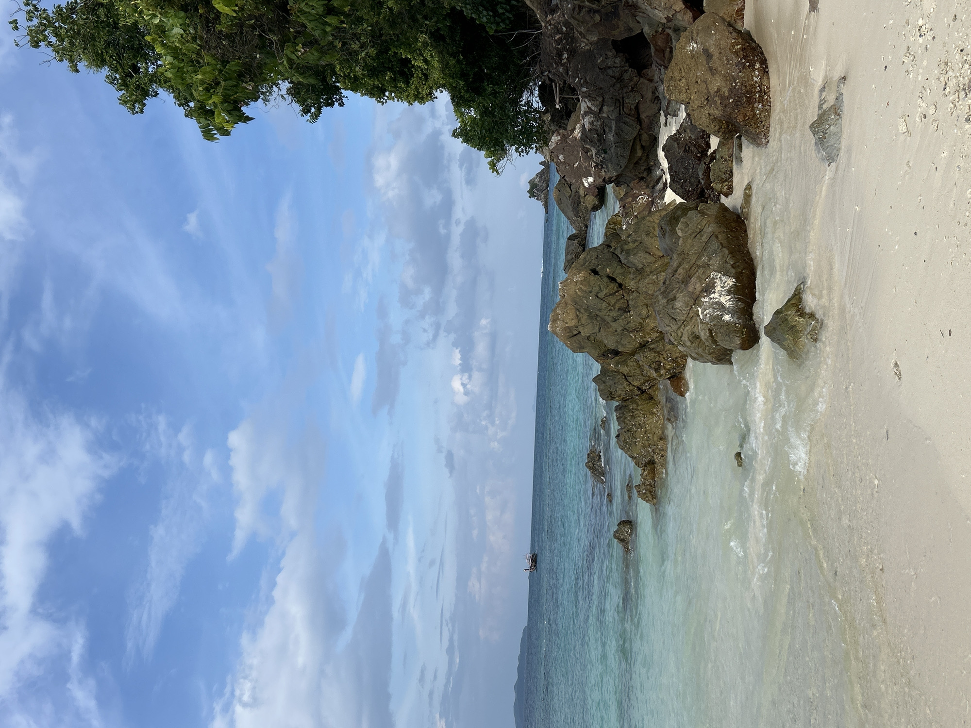 This is an eye-level, wide shot of a tropical beach on Tambon Phru Nai, Thailand. The sand is light in color and appears wet near the water's edge. Large, dark rocks are scattered along the shore, some partially submerged in the clear, turquoise water. The water gently laps at the sand, creating small waves. In the distance, a long-tail boat is visible on the horizon, near a hazy landmass. The sky is a bright blue, with scattered white clouds. Lush green trees and foliage cover a rocky outcrop on the right side of the frame. The overall impression is one of a serene and natural coastal landscape.