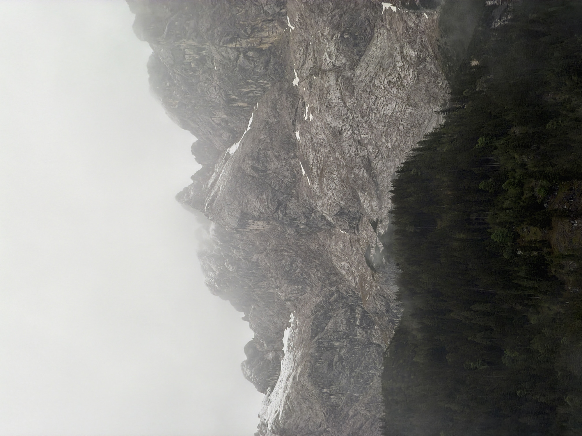 The image displays a dramatic mountain landscape enveloped in mist, likely in the Bavarian Alps near Grainau, Germany. A massive, rugged mountain face, composed of dark grey, heavily textured rock with prominent fissures and crags, dominates the scene. Patches of white snow cling to various ledges and crevices on the mountain, indicating cold temperatures or high elevation.
The upper sections of the mountain are partially obscured by thick, white fog or low-hanging clouds, creating an atmospheric and somewhat mysterious ambiance. The overall lighting is soft and diffuse, characteristic of an overcast day.
Along the base of the mountain, a dense forest of dark green coniferous trees stretches, forming a stark visual contrast with the grey rock. The trees are packed closely, appearing verdant and robust even under the muted light. No people, man-made structures, or discernible text are visible. The scene evokes a sense of wild, untamed nature and imposing grandeur.