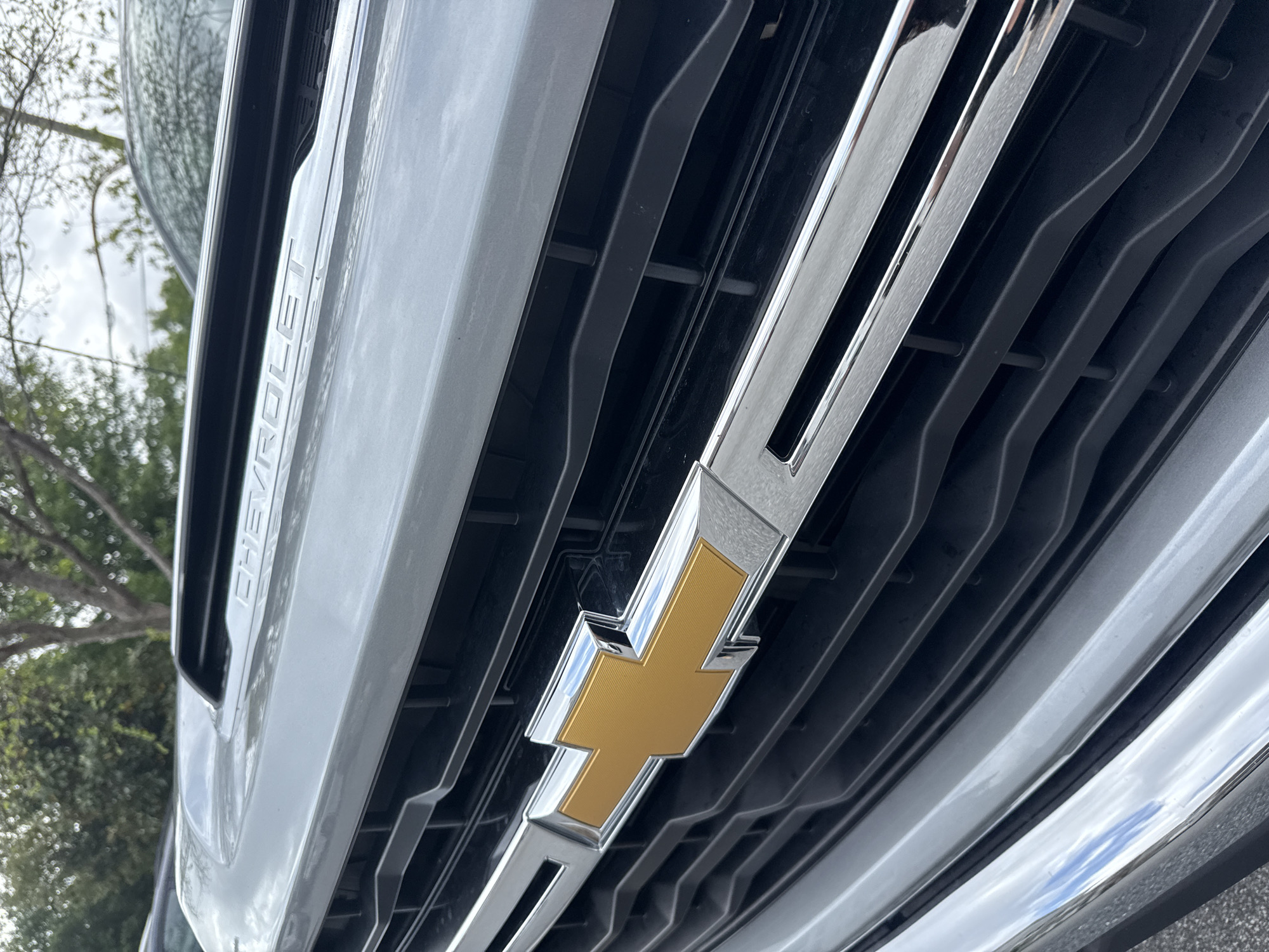 The image shows a close-up of the front grille of a Chevrolet vehicle. The Chevrolet logo, a gold-colored cross or "bow tie" shape outlined in silver or chrome, is prominently displayed in the center of the grille. The grille itself consists of horizontal silver and black slats. The word "CHEVROLET" is visible in silver lettering on the left side of the vehicle's front. The location is suggested to be in Orlando, United States.