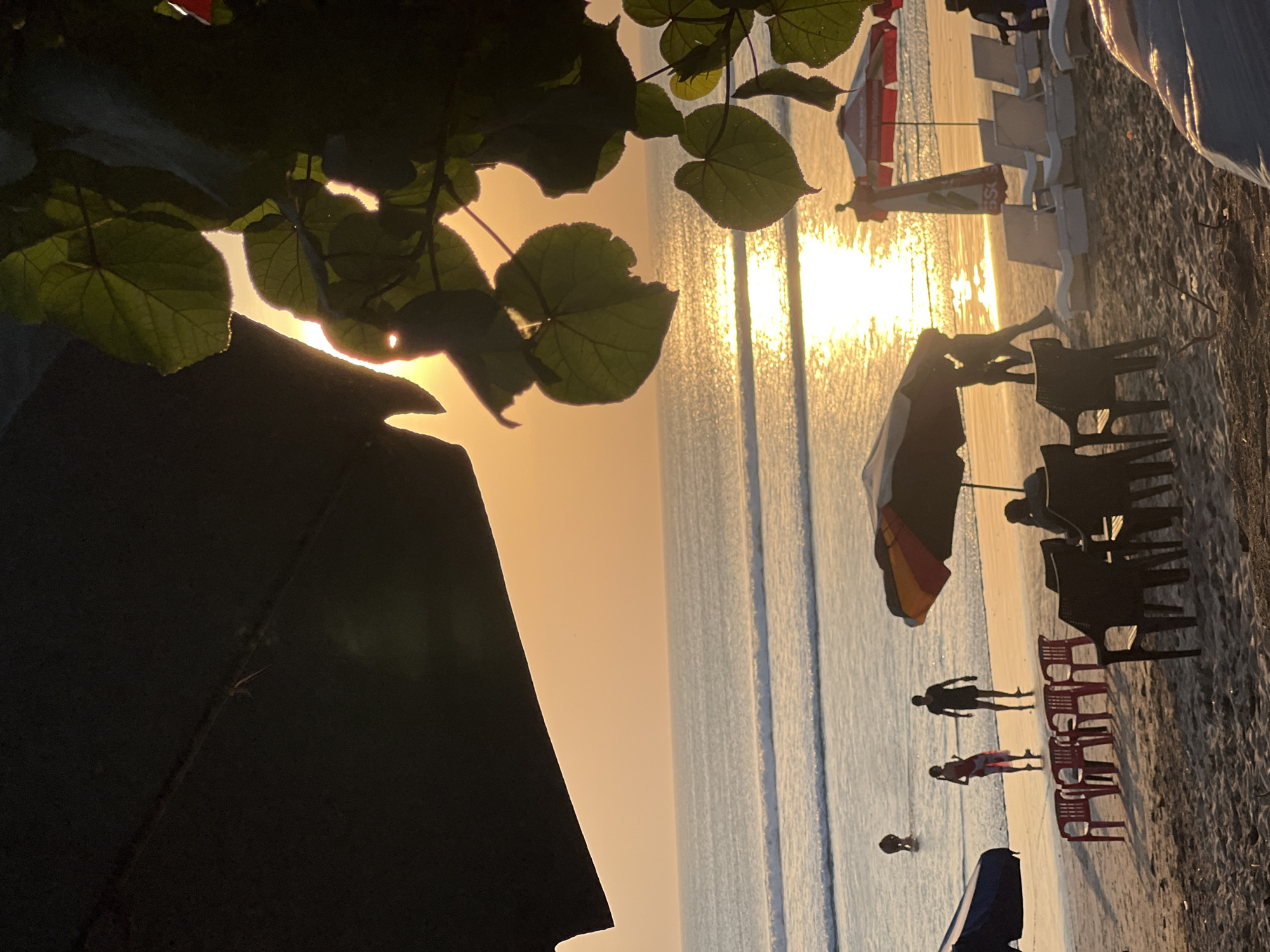 The image depicts a serene beach scene at sunset in Kuta, Indonesia. The golden hues of the setting sun cast a warm glow across the ocean, with gentle waves rolling onto the shore. In the foreground, beach chairs and umbrellas are set up on the sand, some occupied by people relaxing. The ocean is dotted with a few individuals, some wading in the water and others appearing to be walking along the shoreline. Lush green leaves frame the top and right edges of the image, adding a natural element to the composition. The overall atmosphere is tranquil and peaceful, capturing the essence of a perfect evening by the sea in Kuta.