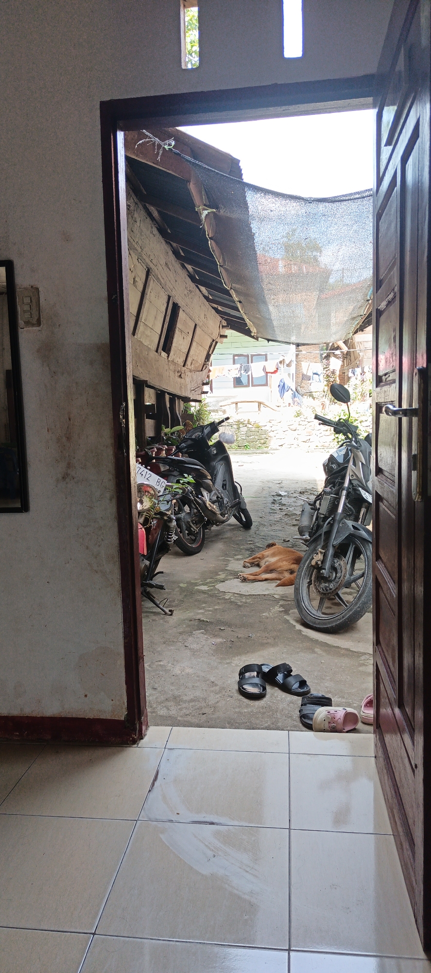The image shows a view through an open doorway into an outdoor area. On the left, part of a white wall with a mirror and an electrical outlet is visible. The doorway leads to a concrete courtyard with two motorcycles parked. One motorcycle is black and red, with the license plate "7412 BG". The other is black and silver. A brown dog is lying down on the concrete between the motorcycles. Scattered on the concrete floor in front of the doorway are several pairs of sandals, including black slides and a pair of pink and white child's shoes. Beyond the motorcycles, a structure with a corrugated metal roof supported by wooden beams is visible, covered in part by a dark mesh. Clothes are hanging on a line in the background. The scene appears to be a residential outdoor space.