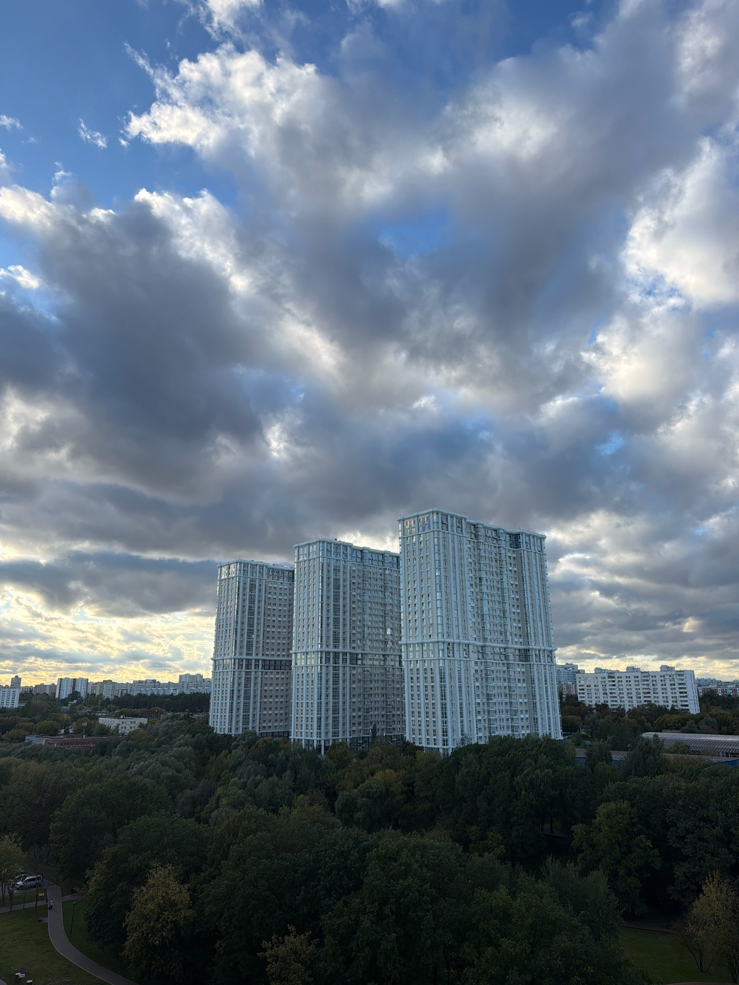 The image captures a scene in Moscow, Russia, featuring a cluster of modern high-rise apartment buildings against a dramatic sky filled with dark, heavy clouds. In the foreground, a dense canopy of dark green trees forms a natural barrier, obscuring the lower portions of the buildings and creating a stark contrast between the natural and urban elements. The trees appear lush and are in full foliage, suggesting a warmer season, likely late summer or early autumn.
The architecture of the apartment buildings is characterized by their pale, light-colored facades and numerous windows, arranged in a repetitive pattern that emphasizes their verticality. They stand in sharp relief against the turbulent sky, which is a mix of deep grays and lighter, illuminated patches, hinting at sunlight breaking through the clouds or the sun setting. The buildings are situated on the outskirts of the city, as indicated by the presence of a park or wooded area in the foreground and a more sprawling, lower-rise urban landscape visible in the distance behind the main cluster of high-rises.
The lower left of the image shows a glimpse of a park area with a paved walkway curving through a grassy expanse. A few cars are parked along a road bordering the park, and a single person can be seen walking on the path, adding a subtle human element to the otherwise imposing urban and natural landscape. The overall impression is one of a developing urban area where nature, represented by the extensive tree cover, is being integrated with or bordered by modern residential developments. The dramatic cloud formations lend a sense of atmosphere and perhaps a touch of melancholy or anticipation to the scene.
