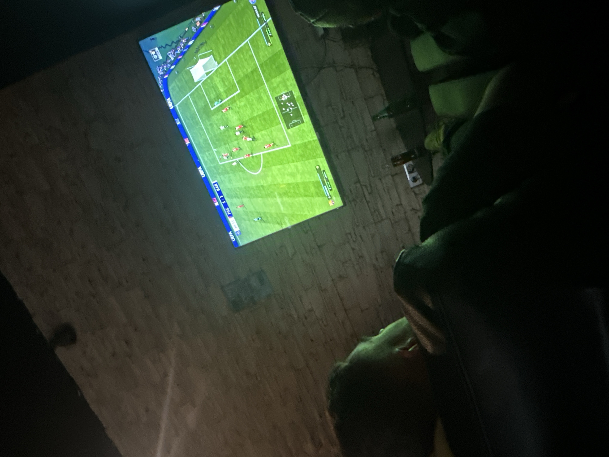 The image depicts a person watching a soccer game on a television. The game appears to be "FIFA." The location is indoors with brick walls. There is a sofa and some bottles visible. There is no information about Plovdiv, Bulgaria in the image.