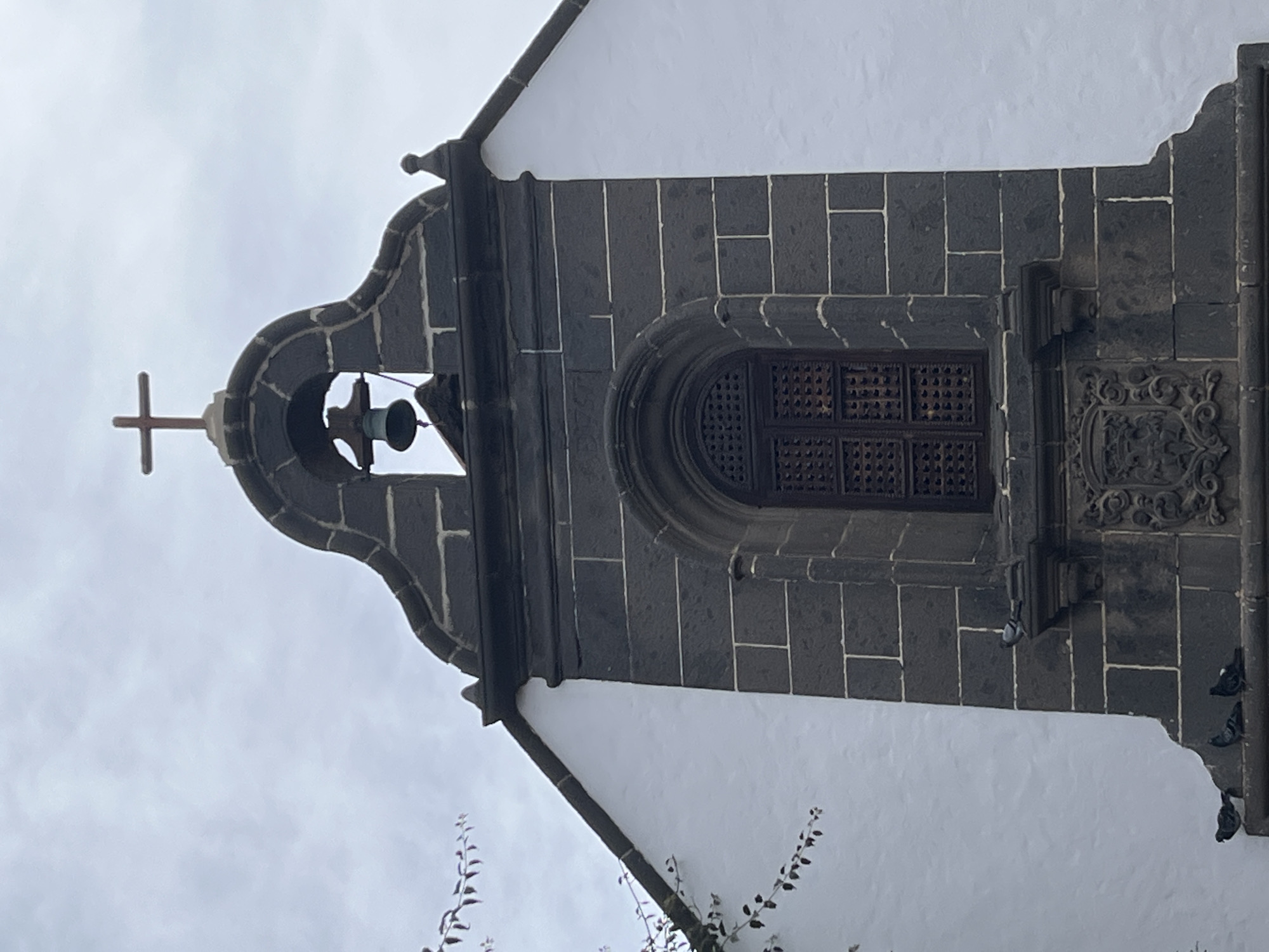 This image shows a partial view of a building facade in Las Palmas De Gran Canaria, Spain. The facade is predominantly white, with architectural details made of dark, possibly volcanic, stone. The stone elements include a bell tower with a cross atop, a window with an arched top and a dark wooden lattice covering, and a decorative carved panel. The tower's design appears to be of a classic architectural style. The bell is visible within the bell tower, and there are a few birds perched on the stone facade at the building's edge. Some branches of a plant are visible at the bottom of the frame. The sky is overcast.