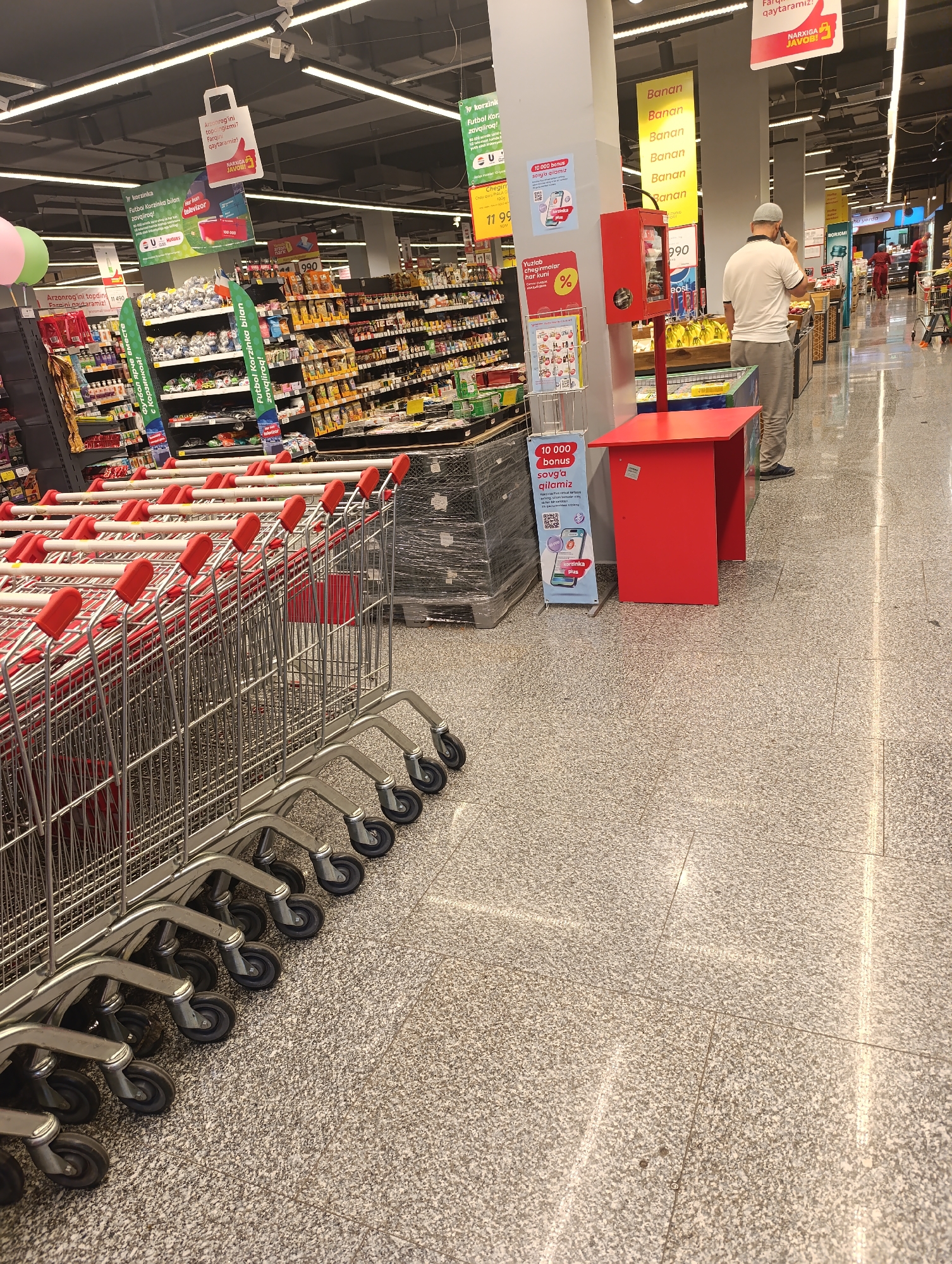 The image captures a row of parked shopping carts in what appears to be a supermarket aisle. The carts are predominantly silver with red handles. In the background, shelves stocked with various products are visible, suggesting a retail environment. A man in a white shirt and grey trousers is standing near a red display unit, holding a phone to his ear. Various promotional signs and posters are displayed on the walls and hanging from the ceiling. The floor is made of speckled grey tiles. The scene is well-lit by overhead fluorescent lighting. The image context indicates this location is in Jizzakh, Uzbekistan.