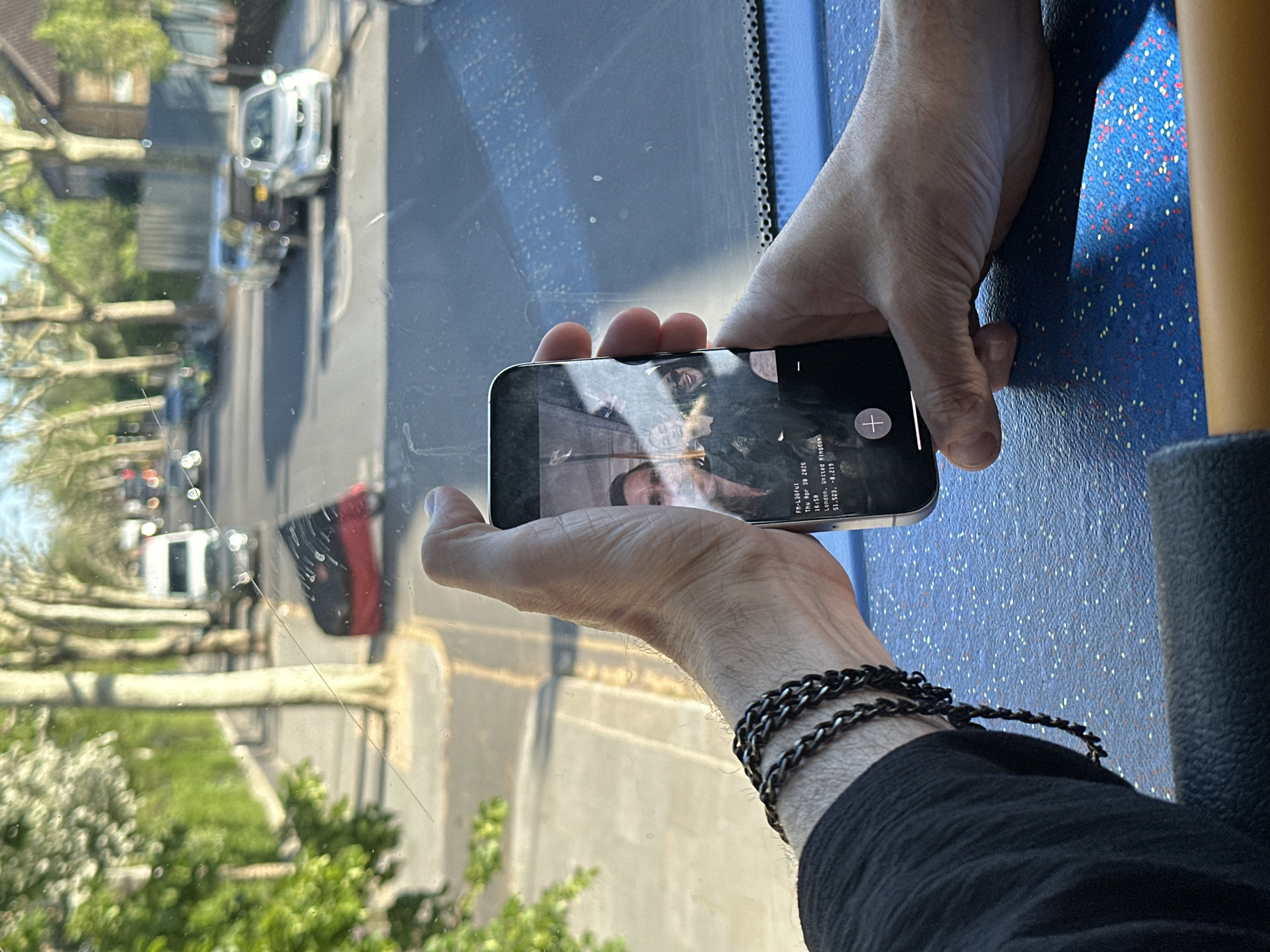 The image, rotated 90 degrees clockwise, presents a close-up view of a person's hands holding a smartphone. The hands are fair-skinned, and the left wrist wears a dark, multi-strand chain bracelet. The individual is dressed in a dark, possibly textured, long-sleeved garment.
The smartphone is held horizontally. Its screen displays what appears to be an image of two people, partially obscured by reflections. Overlaid text on the screen reads: "FM-L36fu1", "Thu Apr 30 2026", "16:50", "London, United Kingdom", and coordinates "51.523, -0.219". A white plus sign icon is also visible.
Through a somewhat dirty window in the background, a bright, sunny urban or suburban street scene is visible. A road runs vertically to the left, lined with trees, some showing new spring foliage. Several vehicles, including a light-colored van and a red car, are present on the street. The clear, bright sunlight indicates it is daytime.
The immediate surroundings within the vehicle feature a blue surface with small white, yellow, and red speckles, typical of public transport seating (likely a bus seat), and a dark grey textured surface, possibly a window frame or part of a seat. The overall scene captures a moment inside a vehicle, looking out onto a London street.