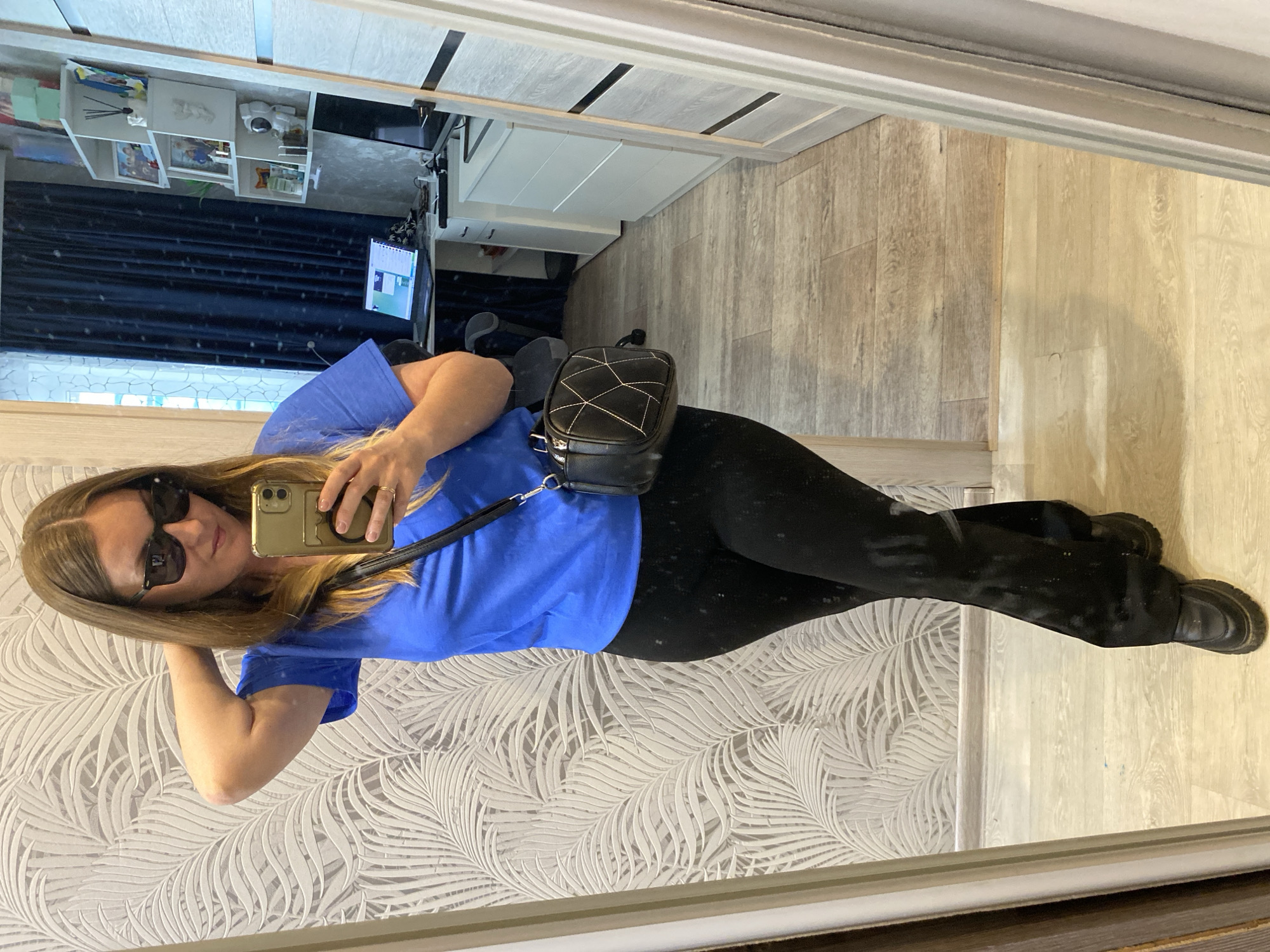 A woman is taking a selfie in a mirror inside an apartment in Noviy, Russia. She is wearing sunglasses, a blue t-shirt, and black pants, with a small black purse slung over her shoulder. The apartment has wood-look floors and a light-colored wall with a tropical leaf pattern. In the background, there is a white cabinet with items displayed on shelves, and a TV on a wall mount. The scene suggests a casual, personal moment captured within a domestic setting.