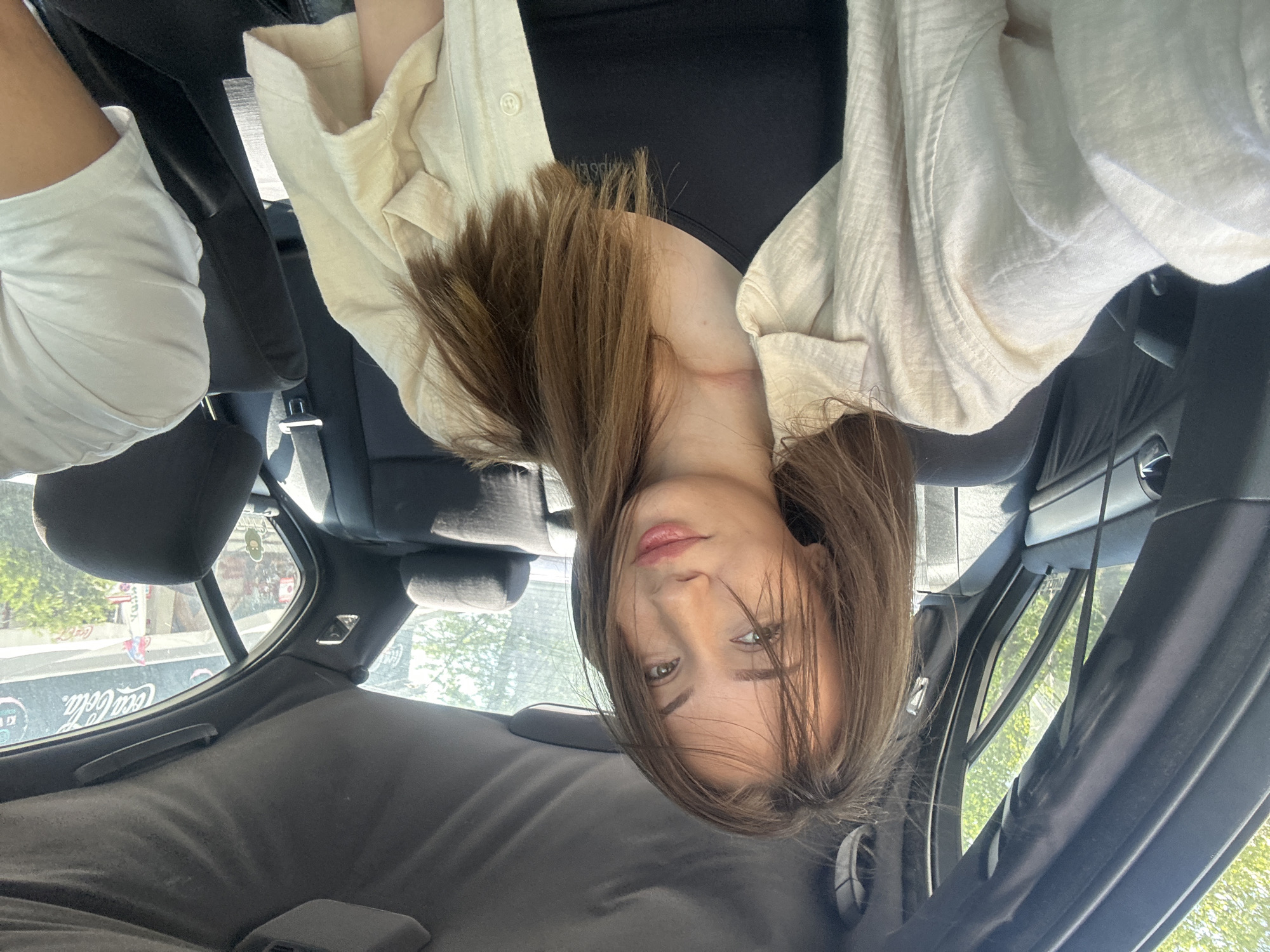 A young woman with long brown hair, wearing a cream-colored blouse and a black top, is taking a selfie inside a car. The car's interior is gray. Outside the car window, the Coca-Cola sign is visible in Burgas, Bulgaria.