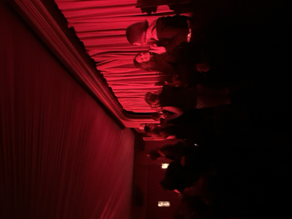 The image depicts an indoor entertainment venue or social gathering space in Paris, France. The scene is illuminated by strong red lighting, casting a deep crimson hue across the entire room. The ceiling is draped with fabric, creating visible folds and lines, leading down to a wall on the right featuring prominent, floor-to-ceiling red velvet curtains. A decorative architectural molding or cornice runs along the top of the wall, above the curtains. On the far left wall, faint, rectangular light fixtures are visible.
Approximately 8 to 10 individuals, appearing to be adults, are present in various parts of the frame, mostly silhouetted against the red light. In the mid-ground, center-right, two women are partially illuminated and engaged in interaction. One woman, with dark, medium-length hair, is seen smiling or laughing as she looks towards the other. The second woman, with short dark hair, has her profile visible and appears to be speaking or listening. She is wearing a dark top with a notable metallic or sequined belt. Other individuals are less distinct, appearing as dark silhouettes standing and mingling in the foreground and background, performing no discernible specific actions. The overall atmosphere suggests a social event or a lively evening gathering within a club, lounge, or cabaret setting. The specific name or address of the venue is not discernible from the visual information provided.