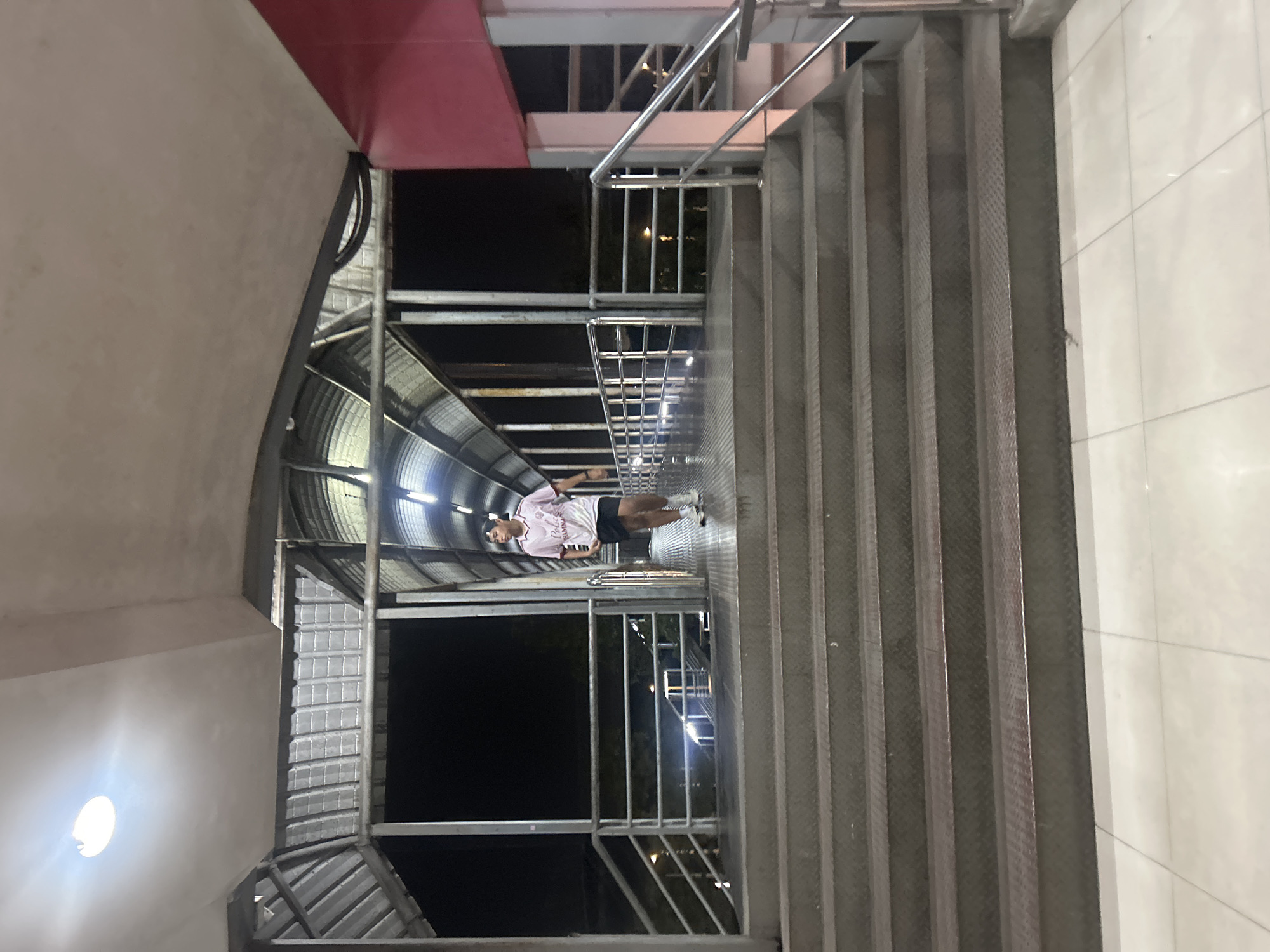 A person stands on a metal staircase with a checkered pattern. The person is wearing a pink t-shirt with white lettering and black shorts. They are looking towards the left and appear to be posing for the photo. The staircase is part of an elevated walkway or overpass. The scene is dimly lit, suggesting it is nighttime. In the background, there are more metal structures, likely part of the same overpass, with curved roofs and lighting. To the right of the staircase, a white tiled wall is visible.
The provided location context is Khlong Luang Town Municipality, Thailand. The image captures an architectural structure, likely a pedestrian overpass, designed with industrial materials. The lighting suggests an urban setting at night. The overall mood is neutral, with the focus on the person and the surrounding environment.
