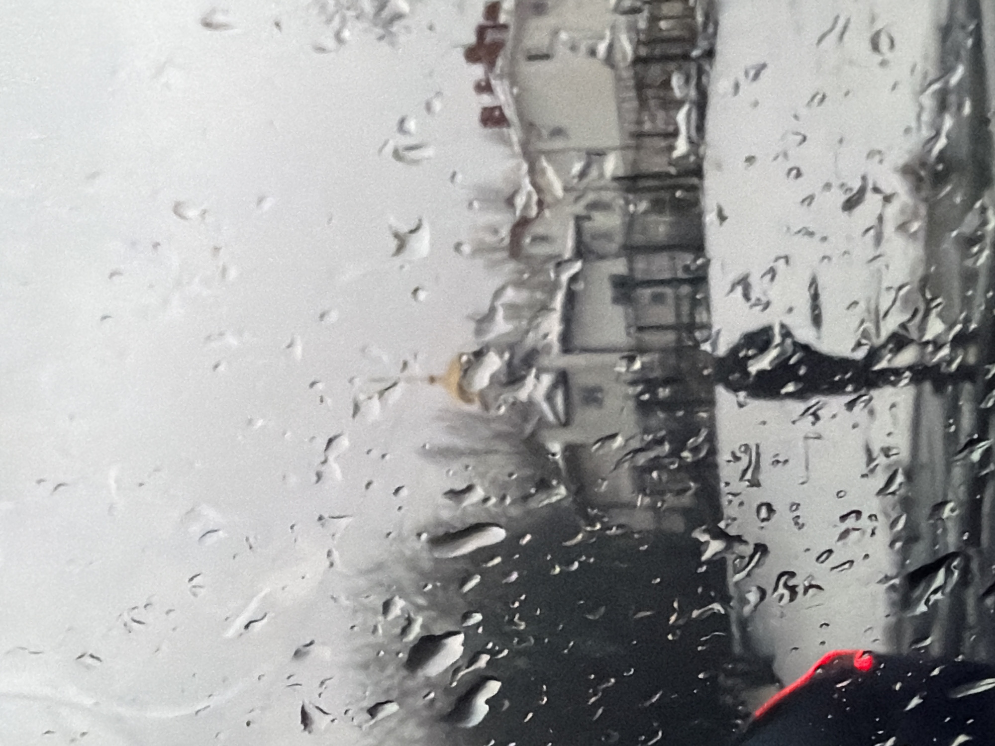 The image captures a blurry and impressionistic view of a cityscape in Moscow, Russia, seen through a window heavily speckled with raindrops. The photograph appears to have been taken from within a vehicle or building, looking outwards during rainy weather.
In the foreground, numerous clear and distinct water droplets of various sizes cover the glass surface, distorting and diffusing the background elements. These raindrops create a textured, shimmering effect across the entire frame.
Beyond the wet glass, the scene consists of indistinct buildings under what appears to be an overcast or gray sky. A prominent feature in the mid-ground is a building with a distinctive golden dome, characteristic of Orthodox churches often found in Russia. Other architectural structures are visible as blurred, blocky shapes, some appearing light gray or white, others darker, suggesting multi-story buildings. To the right, a large, dark, vertical structure, possibly a pillar, another building, or part of an overpass, occupies a significant portion of the frame.
In the lower right corner, a bright red light stands out against the muted tones of the background, likely a car's taillight or a reflection from an interior element. The weather is clearly rainy, indicated by the prominent raindrops, which contribute to the soft, diffused lighting throughout the scene. The time of day appears to be daytime, but with heavy cloud cover. No discernible text is visible in the image.