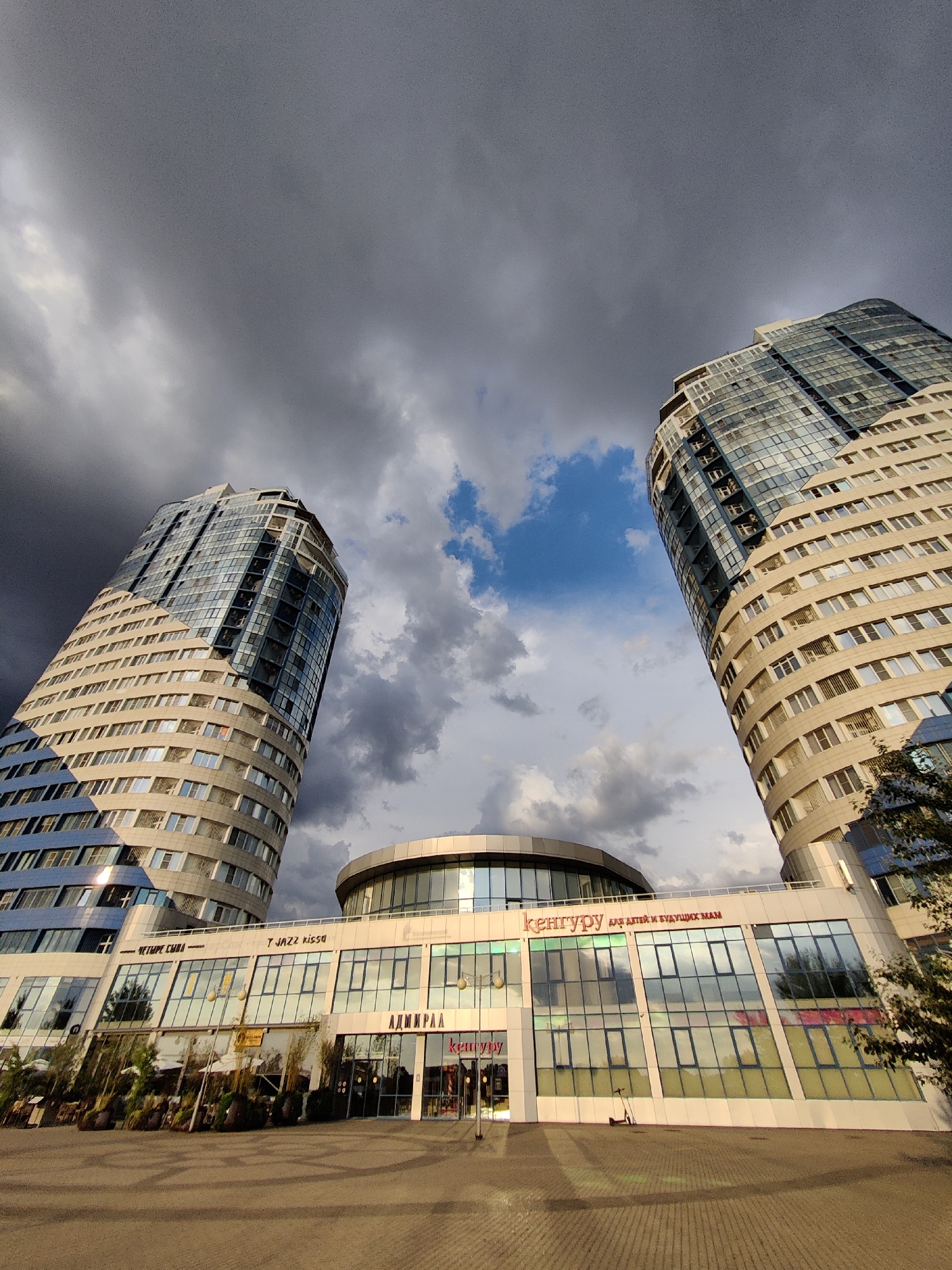 The image captures a wide-angle view of two modern, curving skyscrapers against a dramatic sky. The buildings are tall and sleek, with many windows reflecting the cloudy sky. The lower levels of the buildings feature a commercial strip with large glass facades.
Visible text on the commercial strip includes "T JAZZ kissa", "АДМИРАЛ", and "кенгуру ДЛЯ ДЕТЕЙ И БУДУЩИХ МАМ" (which translates to "Kangaroo For Children And Future Moms"). The overall atmosphere is somewhat moody due to the dark, stormy clouds contrasting with patches of bright blue sky. The lighting suggests it is late afternoon or early evening. The paved ground in front of the buildings indicates an urban outdoor setting, possibly a plaza or walkway. There are no people clearly visible in the image, but the presence of shops suggests a public space.