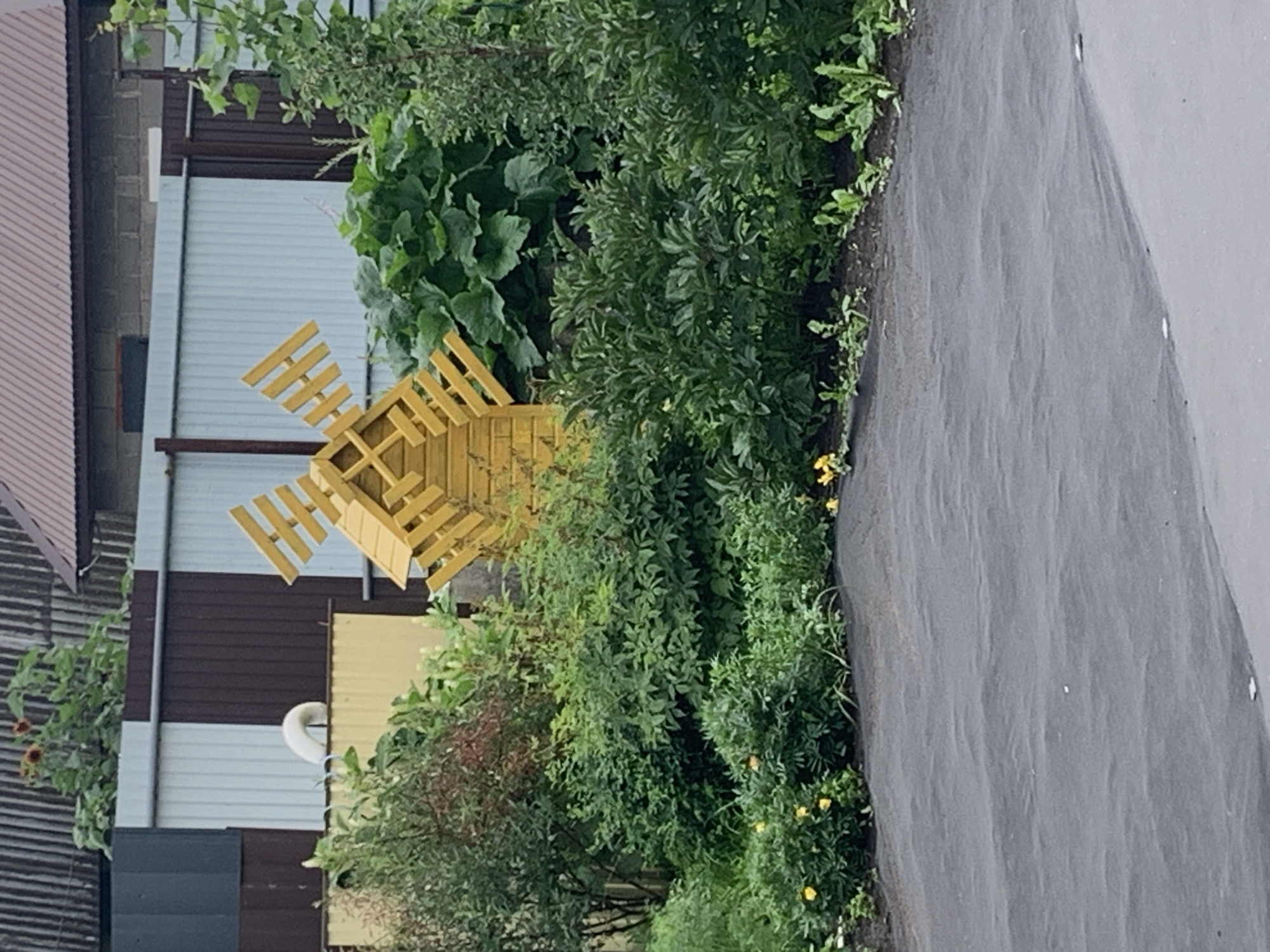 The image depicts a decorative wooden windmill, painted yellow, positioned in a garden area. The windmill is surrounded by lush green bushes and some yellow flowers. In the background, a corrugated metal fence, a brick wall, and parts of a building with a brown roof are visible. A section of dark gray ground cover is present in the foreground. The scene appears to be a residential or garden setting, likely in Dubnik, Russia, given the architectural styles and vegetation. There are no people or animals present in the image, and no actions or interactions are taking place.