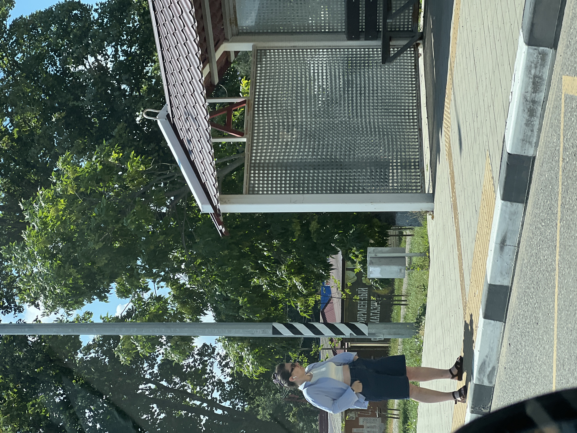 The image captures a street scene in Belozyorny, Russia, featuring a woman standing on a sidewalk next to a bus stop. The woman, wearing a light blue unbuttoned shirt, a white crop top, and dark shorts, has her hands in her pockets. She is also wearing sunglasses and has dark hair pulled back. The bus stop has a brown tiled roof, white pillars, and a perforated metal wall. A wooden bench is visible under the bus stop shelter. The sidewalk is paved with light gray square tiles and bordered by a tactile paving system. A striped pole with black and white bands stands to the left of the woman. In the background, trees with lush green foliage are visible, along with buildings and a sign with Russian text. The overall environment suggests a sunny day in an urban or semi-urban setting.
