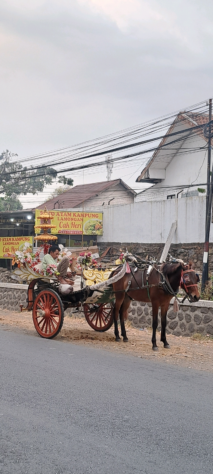 A horse-drawn carriage with two occupants is parked on the side of a paved road in Jogosari, Indonesia. The carriage is decorated with colorful flowers and has red spoked wheels. A man wearing a light green shirt and brown pants sits in the carriage, looking down. Another person, partially obscured, is seated beside him. The horse, a dark brown mare with a bridle and harness, stands facing right, tethered to the carriage. Behind the carriage, a white wall with a stone foundation runs along the property line, with buildings and overhead electrical wires visible in the background. A yellow sign with text in Indonesian and an image of food is displayed on the left. The ground is a mix of asphalt and dirt with some dry vegetation.