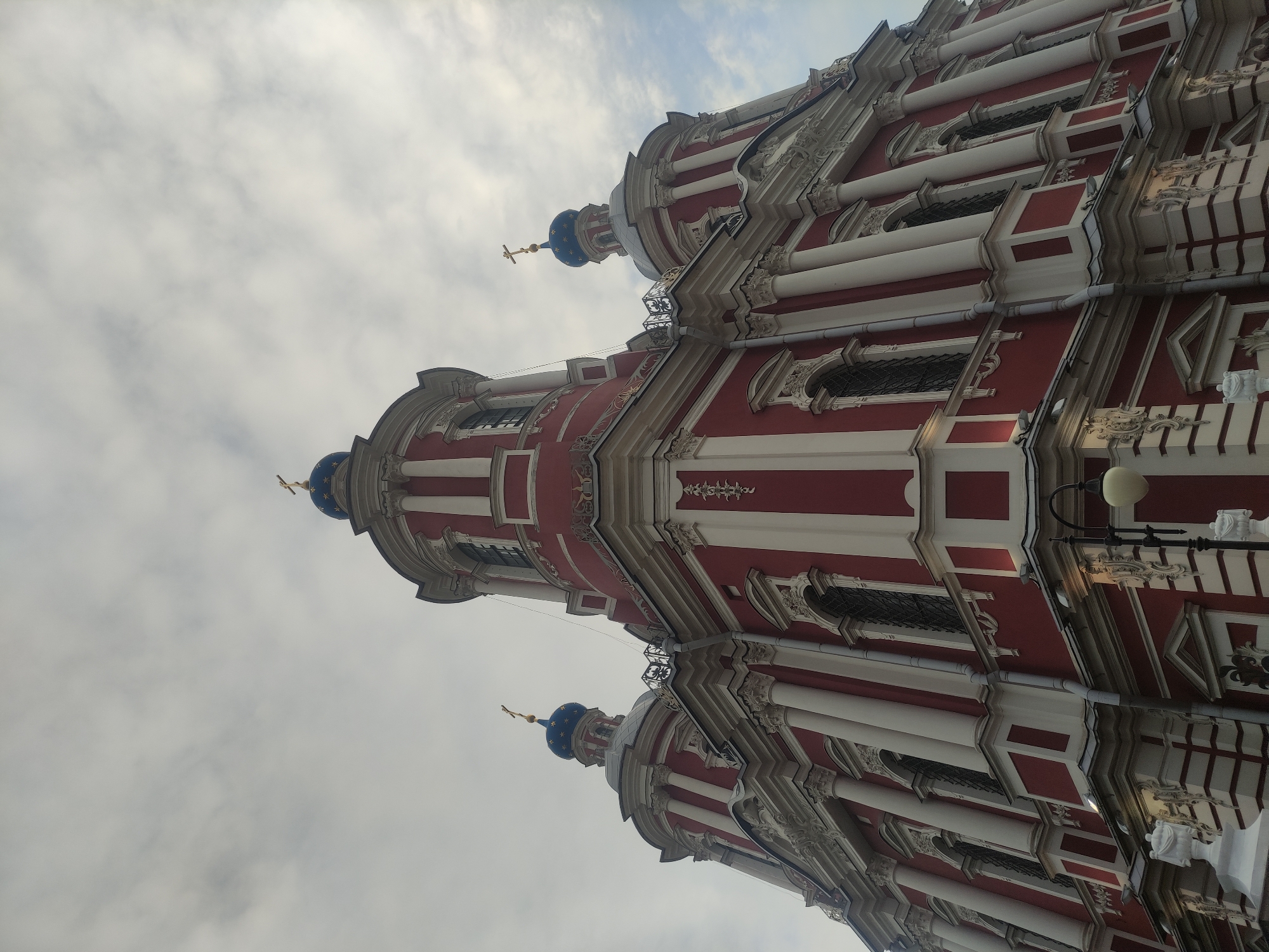 The image depicts a building in Moscow, Russia. The building is red and white, with multiple tiers and a dome topped with a golden cross. The architecture is elaborate, with intricate details and columns. The sky is overcast, suggesting a cloudy day. A lamppost is visible on the right side of the image.