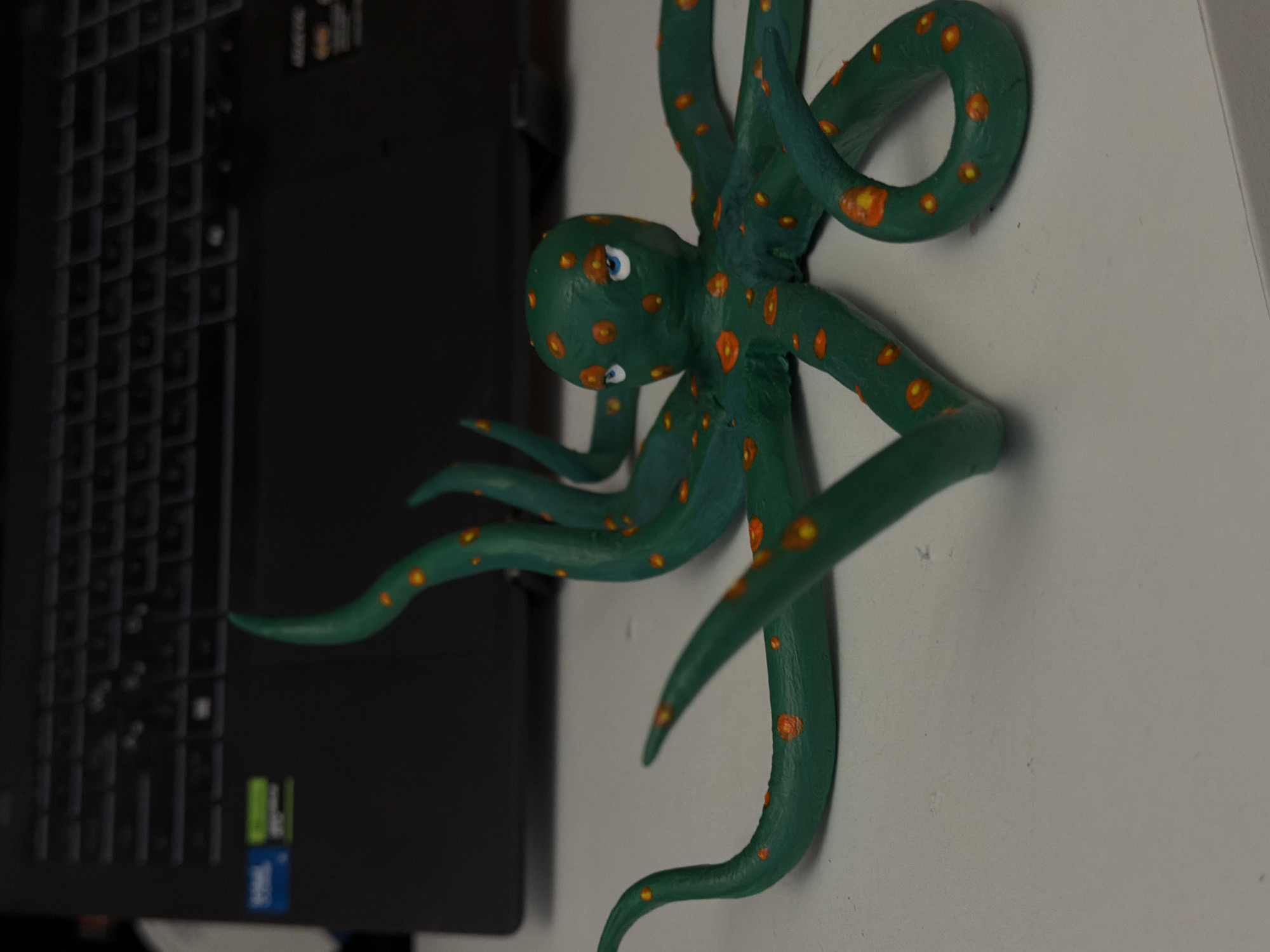 The image, taken in Perm, Russia, features a vibrant green octopus figurine with numerous orange spots and distinctive blue eyes. The octopus appears to be attached to a light-colored, possibly white, vertical surface, with its sculpted tentacles curled and extending in various directions. To the left of the figurine, a black laptop keyboard is partially visible. On the laptop's body, two prominent logos are discernable: a green and black "NVIDIA GeForce RTX" emblem, and a blue and black "Intel Core i7" badge. The scene depicts an indoor setting, likely a desk or personal workspace. No explicit activity is occurring; it is a still life composition of these objects. The lighting suggests an interior environment, without indicating a specific time of day.