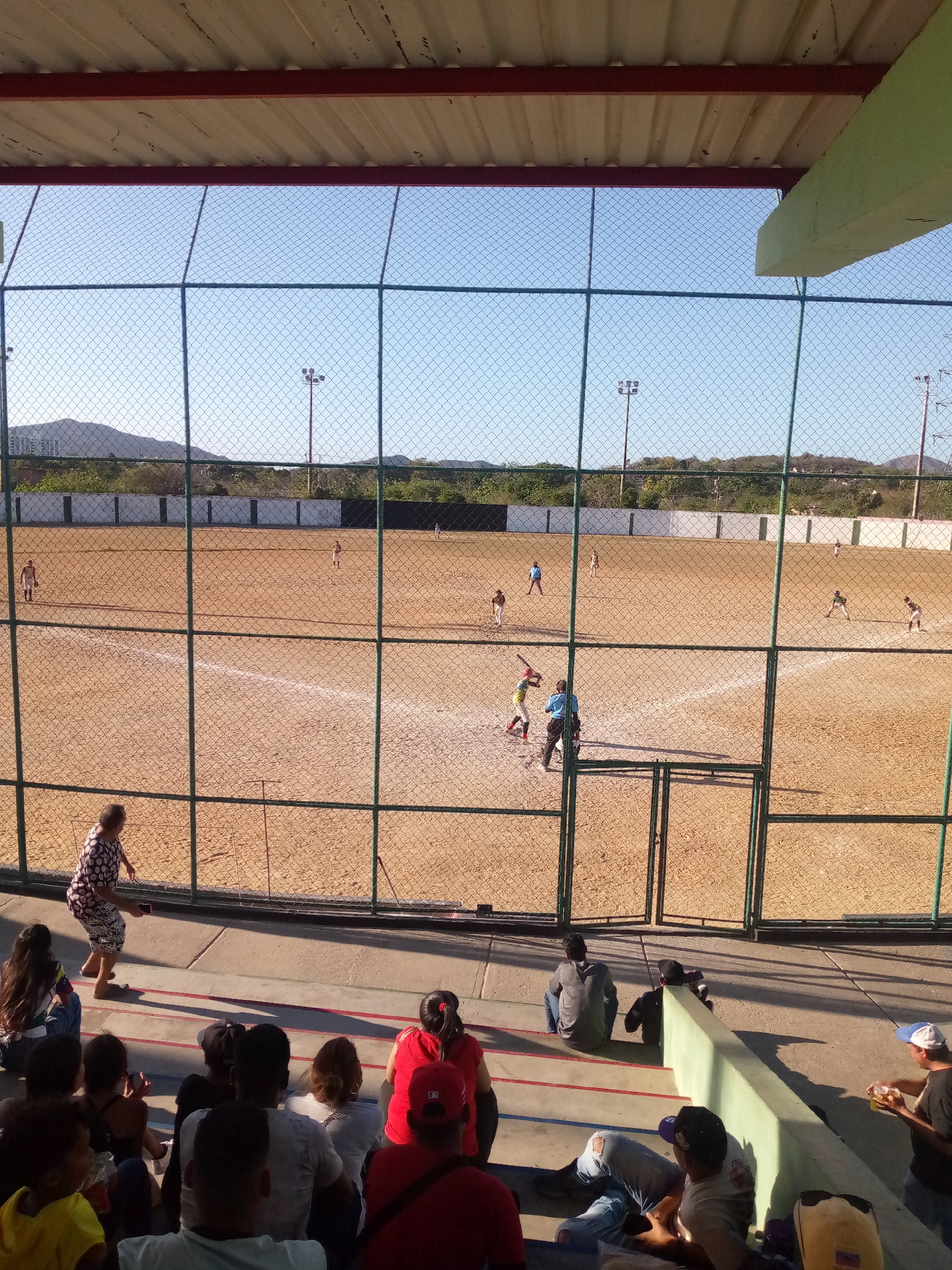 The image shows a baseball game in progress viewed through a chain-link fence from an elevated seating area. The location is Juan Griego, Venezuela, based on visual clues.
In the foreground, spectators are seated on red and grey painted concrete tiers. The spectators are a mix of adults and children. Some are sitting, others are standing. A woman in a patterned dress is walking on the upper tier. Other individuals are sitting or kneeling.
Beyond the fence is a baseball field with several players in action. A batter is at home plate. An umpire is standing nearby. Other players are positioned in the field.
The field is primarily brown dirt, with white lines marking the base paths and batter's box. A low concrete wall with dark green fencing surrounds the field. In the background, a sparse tree line and low hills are visible under a clear blue sky. Two sets of tall light poles are visible.