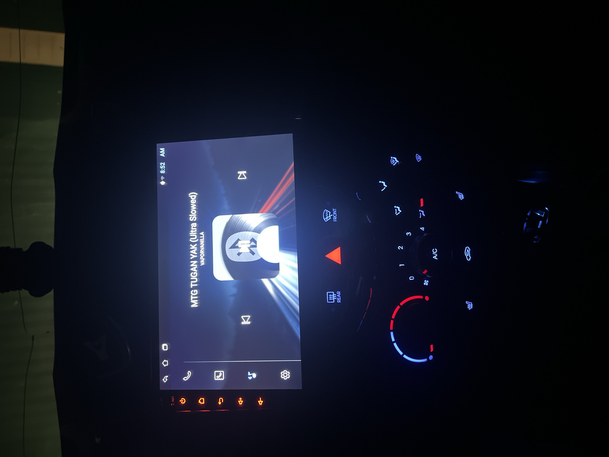 The image shows a car's dashboard at night. The infotainment system displays the song "MTG TUGAN YAK (Ultra Slowed)" by VAPORVANILLA. The climate control panel is illuminated, with settings visible for fan speed, temperature, and air conditioning. The picture appears to be taken in the dark. There is no indication of the location being in the city of Бизяки, Russia.