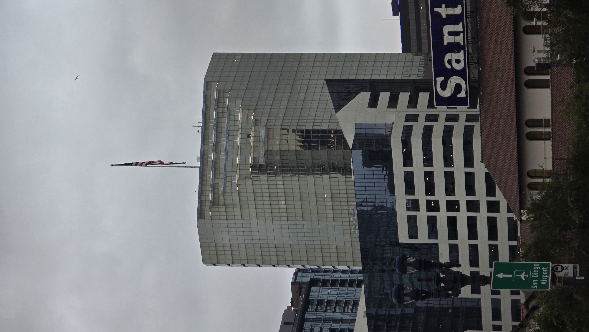 This is a medium shot, eye-level, outdoor, vertical photograph of a cityscape on an overcast day. In the foreground, on the left, there is a black, ornate streetlamp with a dark green sign below it indicating "San Diego Airport" with an airplane symbol and an arrow pointing upwards. To the right of the streetlamp and sign, a red-tiled roof of a building is visible with arched windows. In the mid-ground, there is a multi-story white building with many horizontal windows. Above and behind this building, a large, modern, glass-covered skyscraper dominates the scene. An American flag is flying from a flagpole on top of this skyscraper. A small bird is flying in the sky above and to the right of the skyscraper. To the far right of the image, a large blue billboard with white lettering that appears to read "Sant..." is visible behind the buildings. The sky is a uniform grey color, suggesting cloudy or foggy weather.